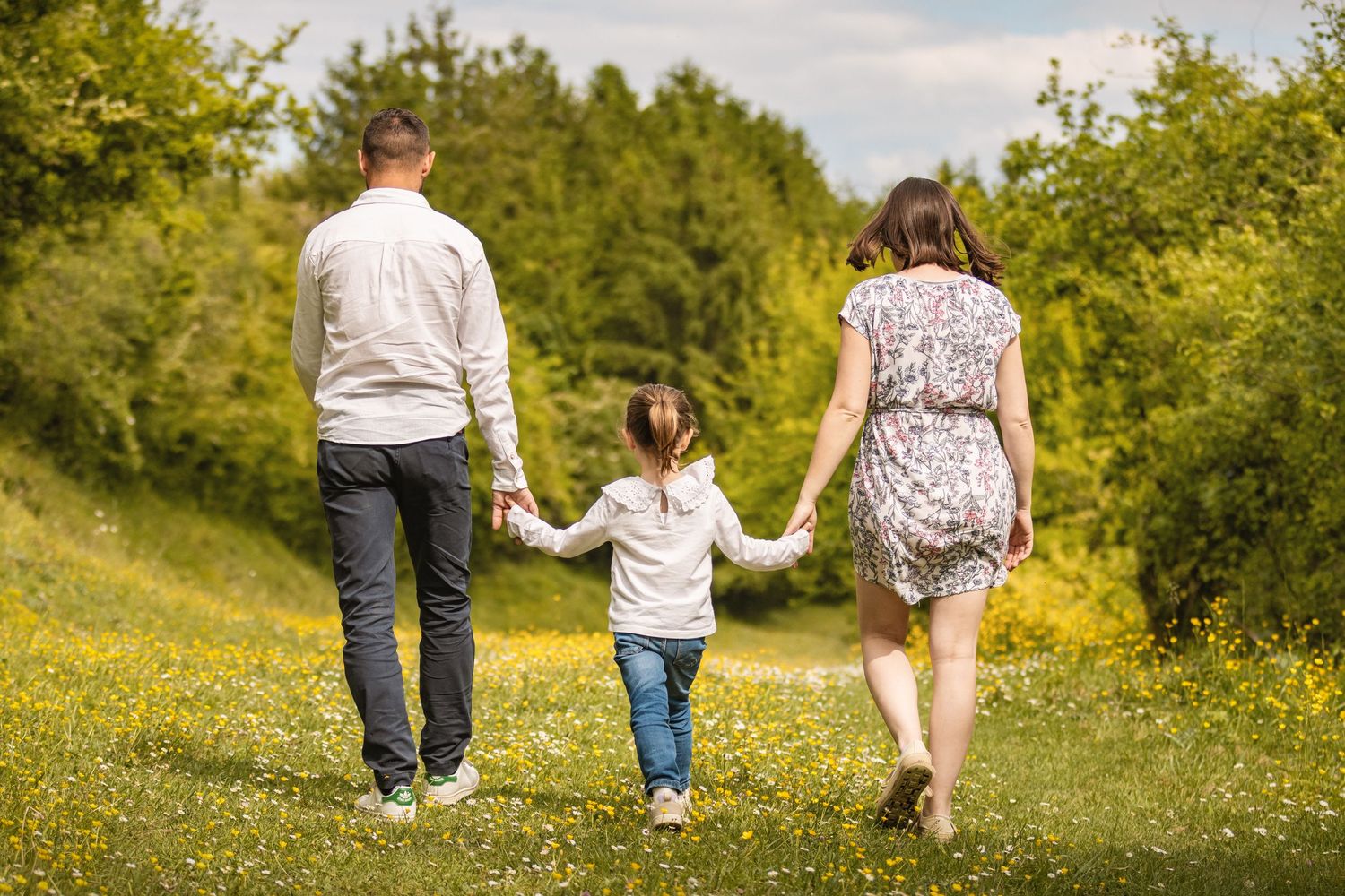 Family of three holding hands walking through a sunny green meadow with wildflowers and trees in background.