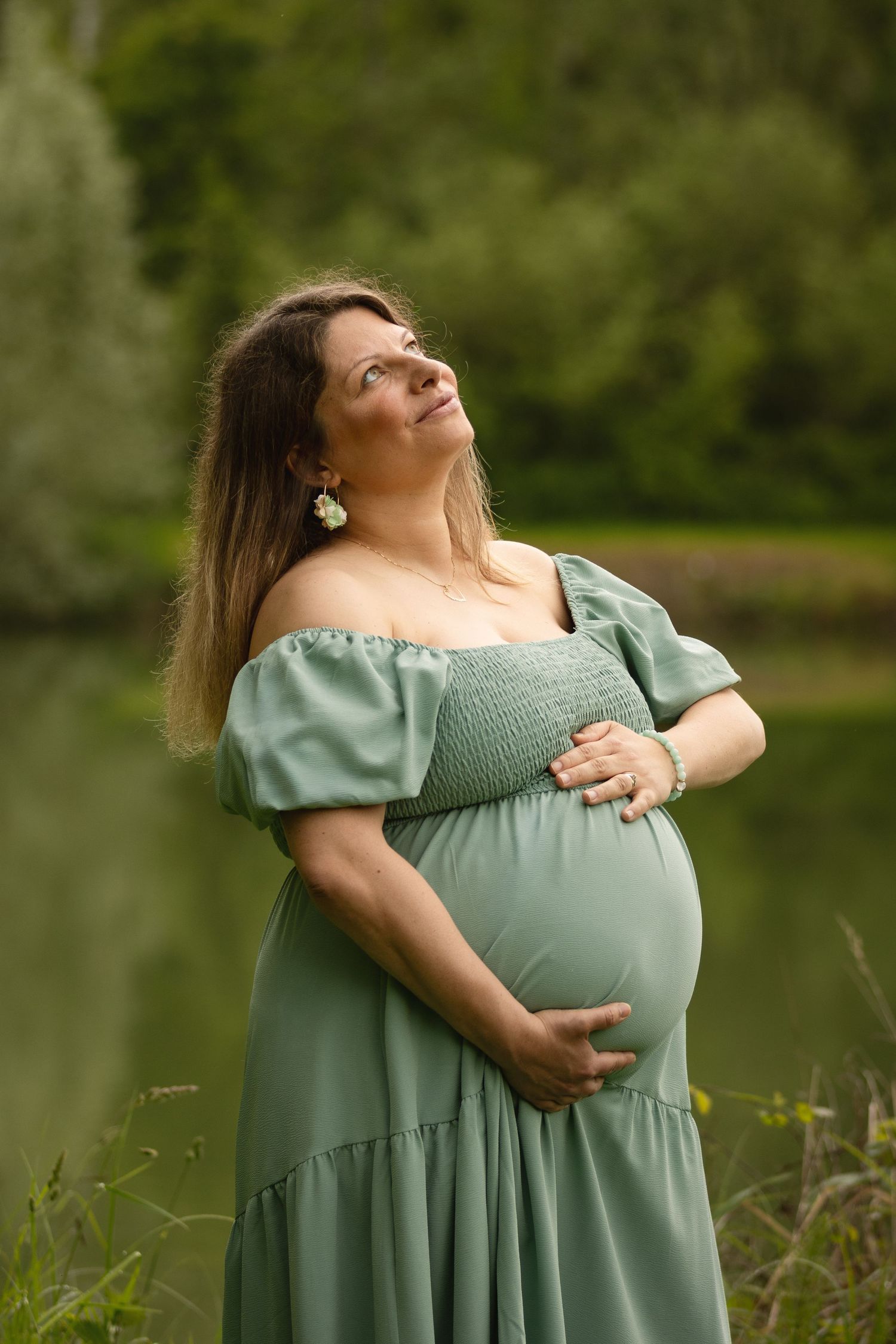 Pregnant woman in sage green off-shoulder dress cradling baby bump outdoors near a serene lake surrounded by lush trees.