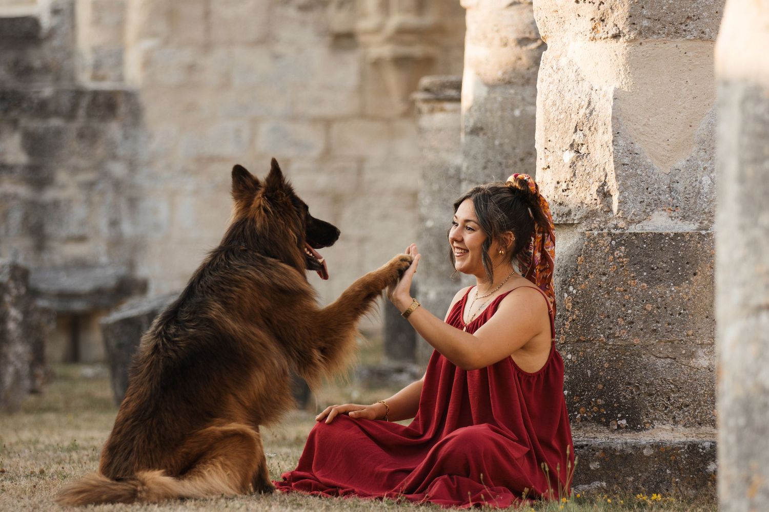Woman in red dress high-fiving her German Shepherd dog near ancient stone ruins.