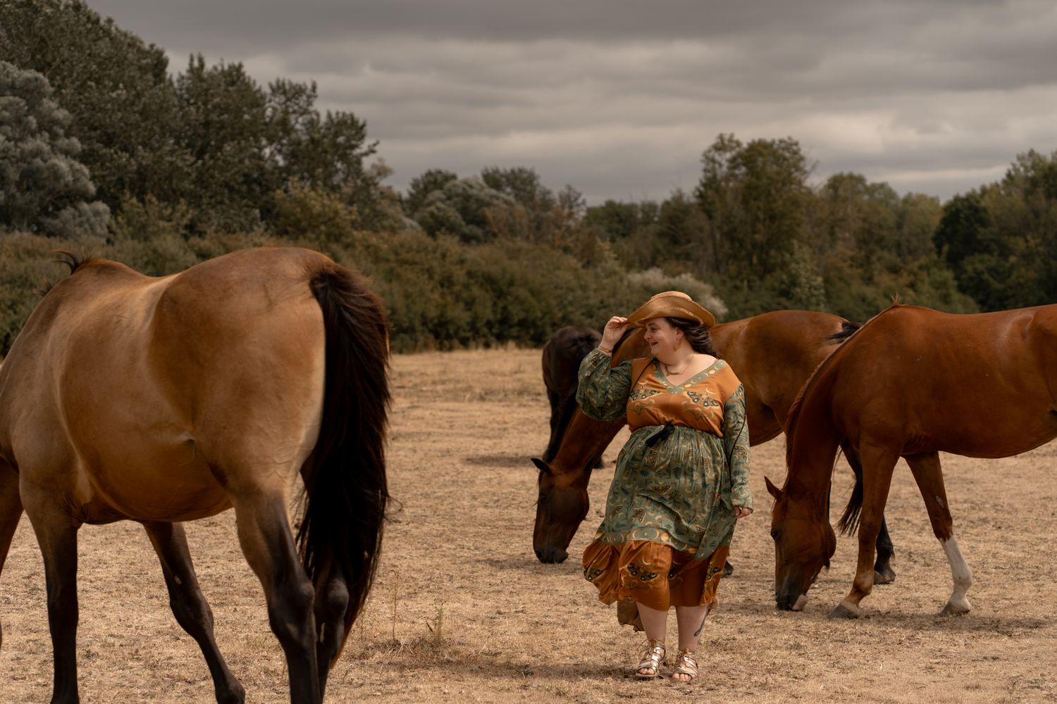 Woman in green dress and hat walking among grazing brown horses in a field.