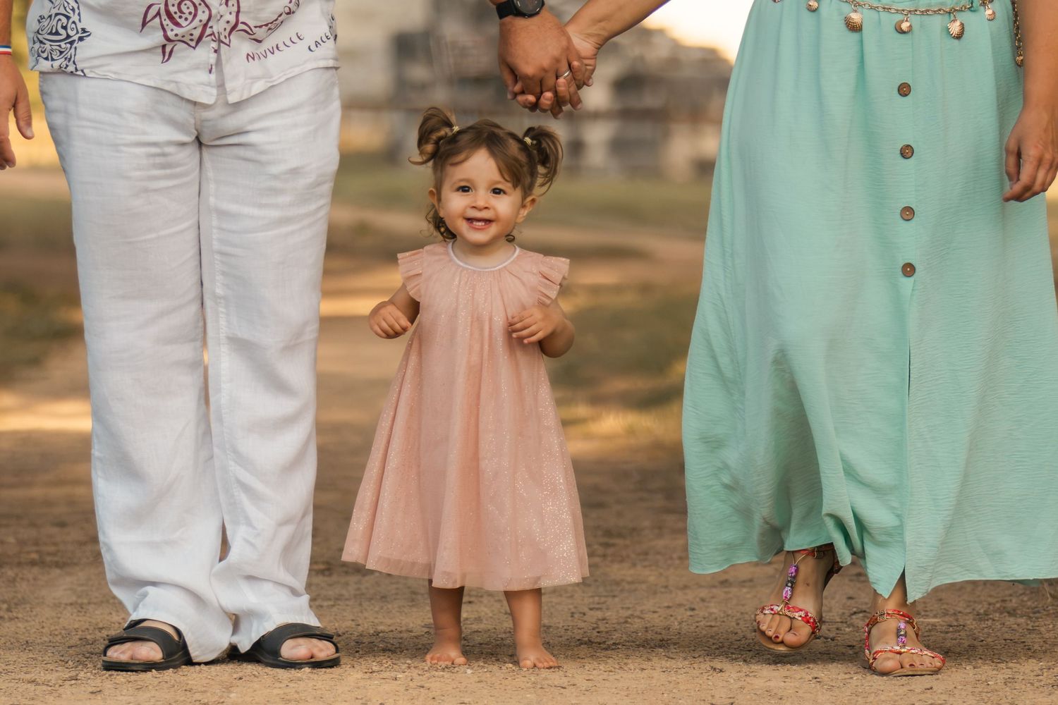 A smiling toddler girl in a pink dress stands barefoot between two adults holding hands outdoors.