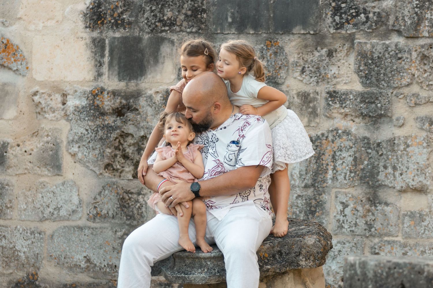A bald father sits on a stone ledge hugging his three young children against an ancient stone wall.