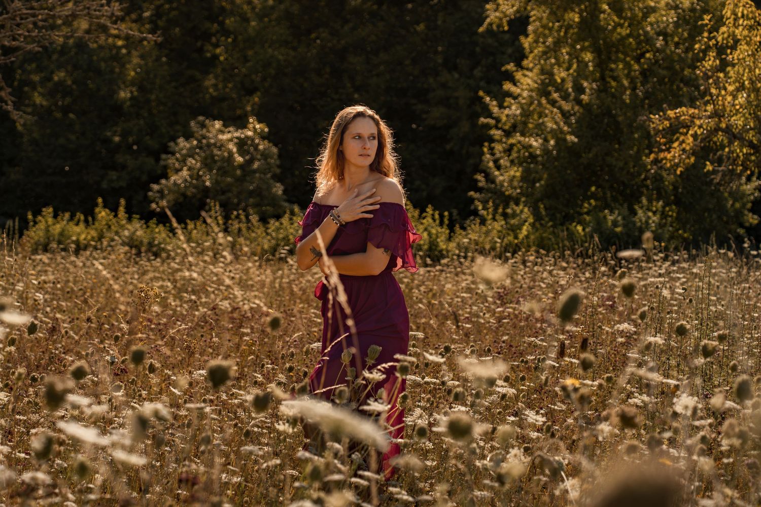 Woman in burgundy dress standing in golden wildflower field with trees in background during golden hour.