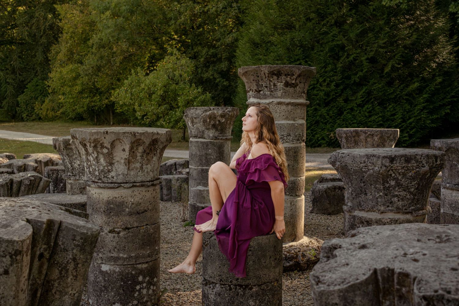 Young woman in purple dress sitting on ancient stone columns surrounded by lush green trees.