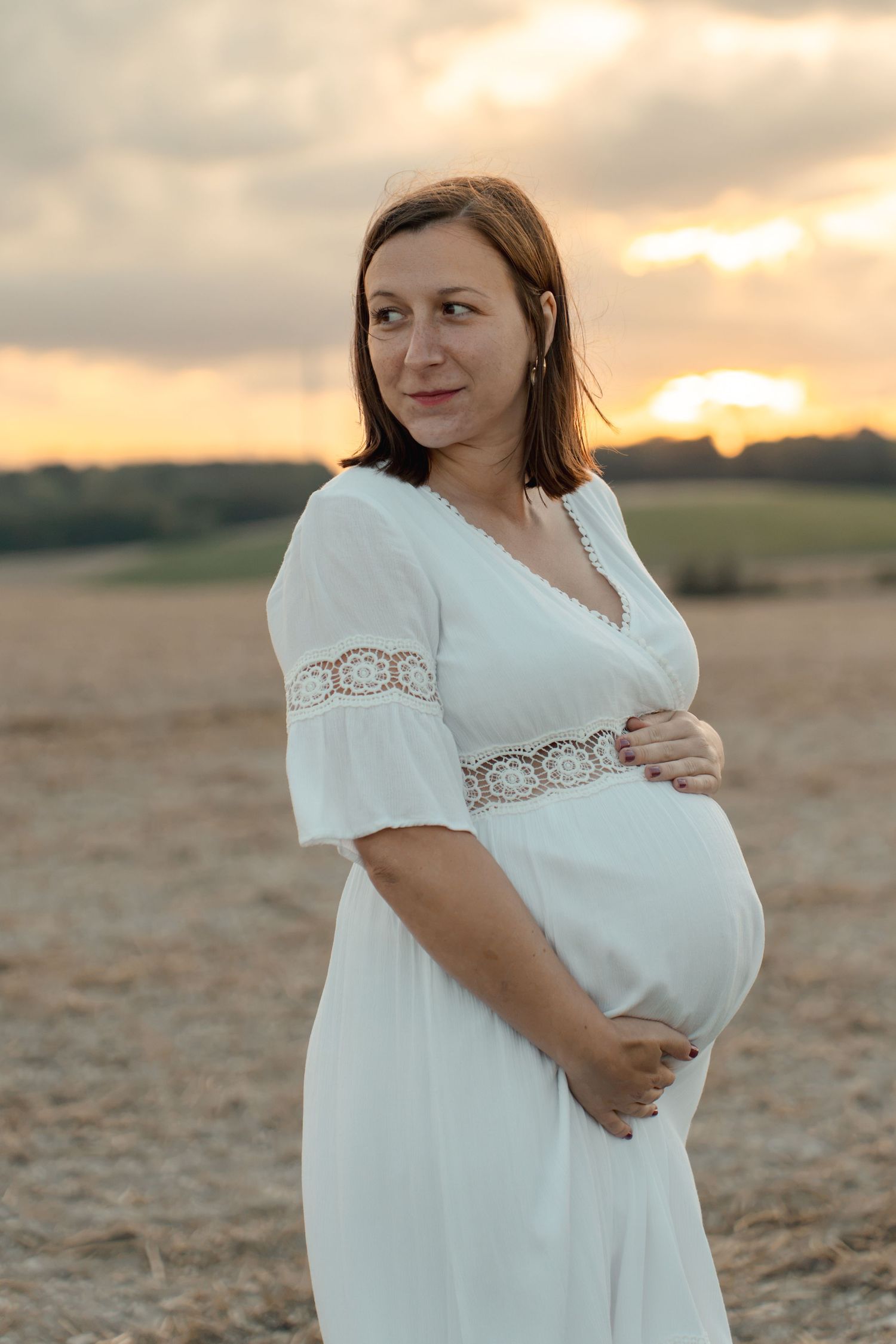 Pregnant woman in white lace dress cradling baby bump in golden field at sunset.