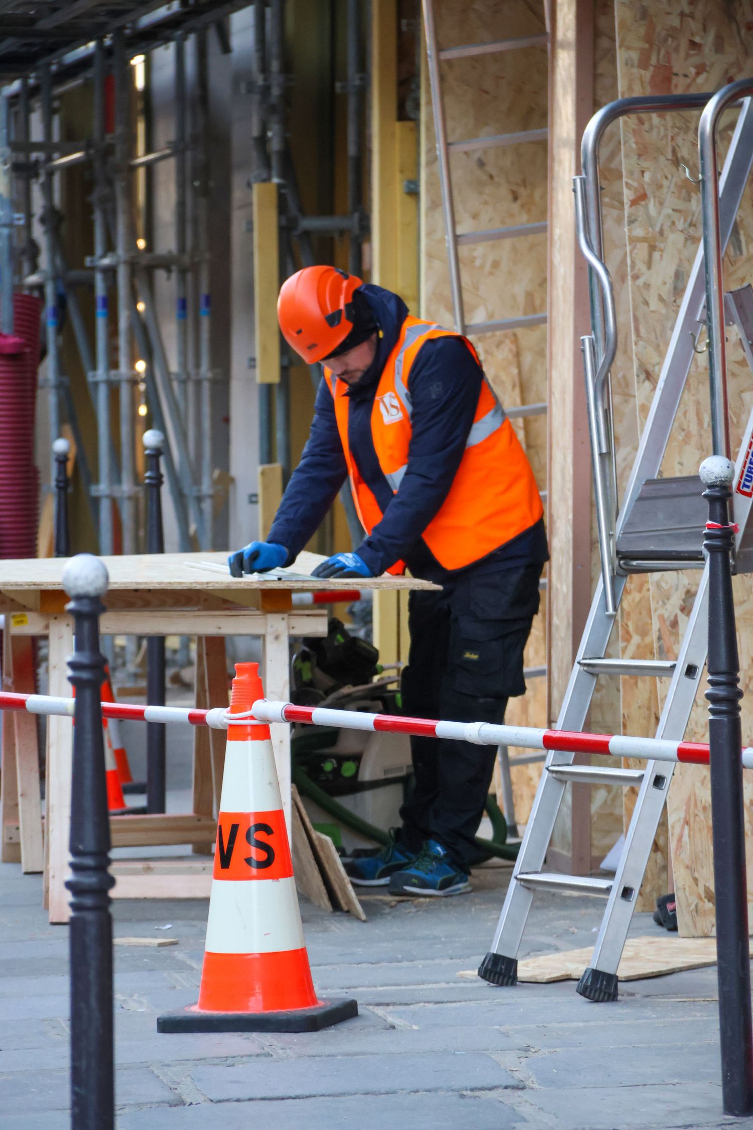 Construction worker in orange safety vest and helmet works with wood planks at an outdoor building site with traffic cones.