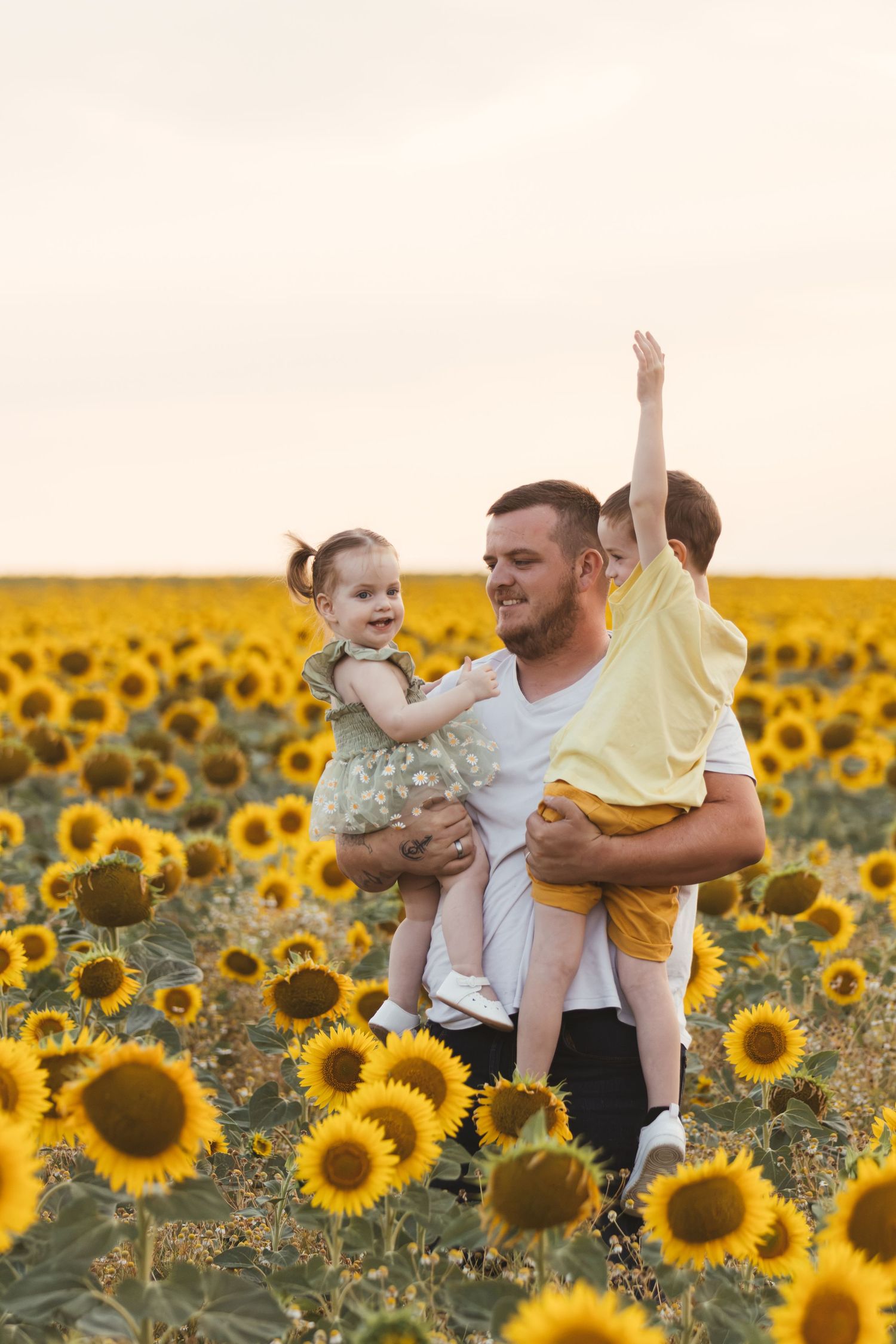 Father holding two young children in a vibrant sunflower field at sunset, with boy raising his arm joyfully.
