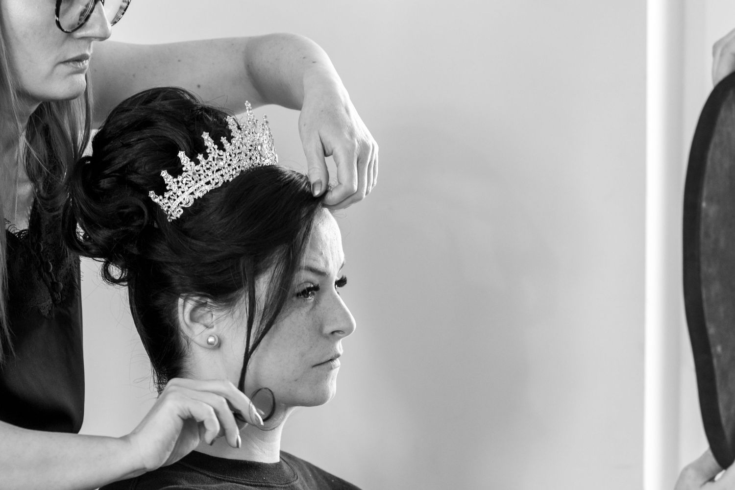 Black and white photo of a hairstylist placing a crystal tiara on a brunette bride's elegant updo.