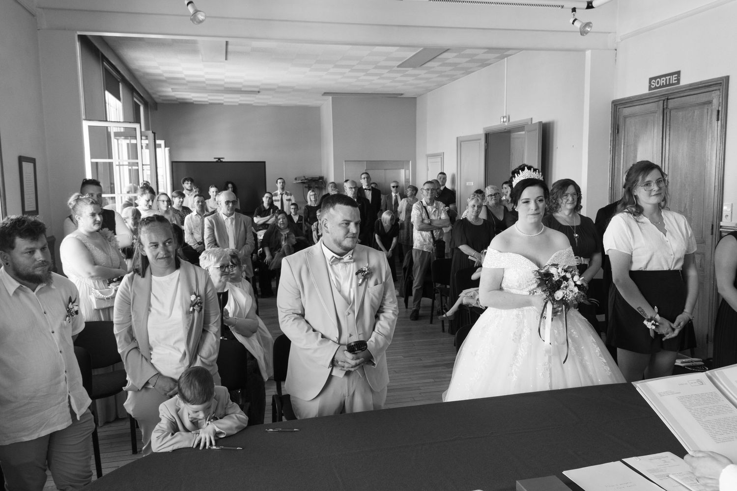 Black and white wedding ceremony photo showing bride in white gown holding bouquet with groom and guests in elegant hall.