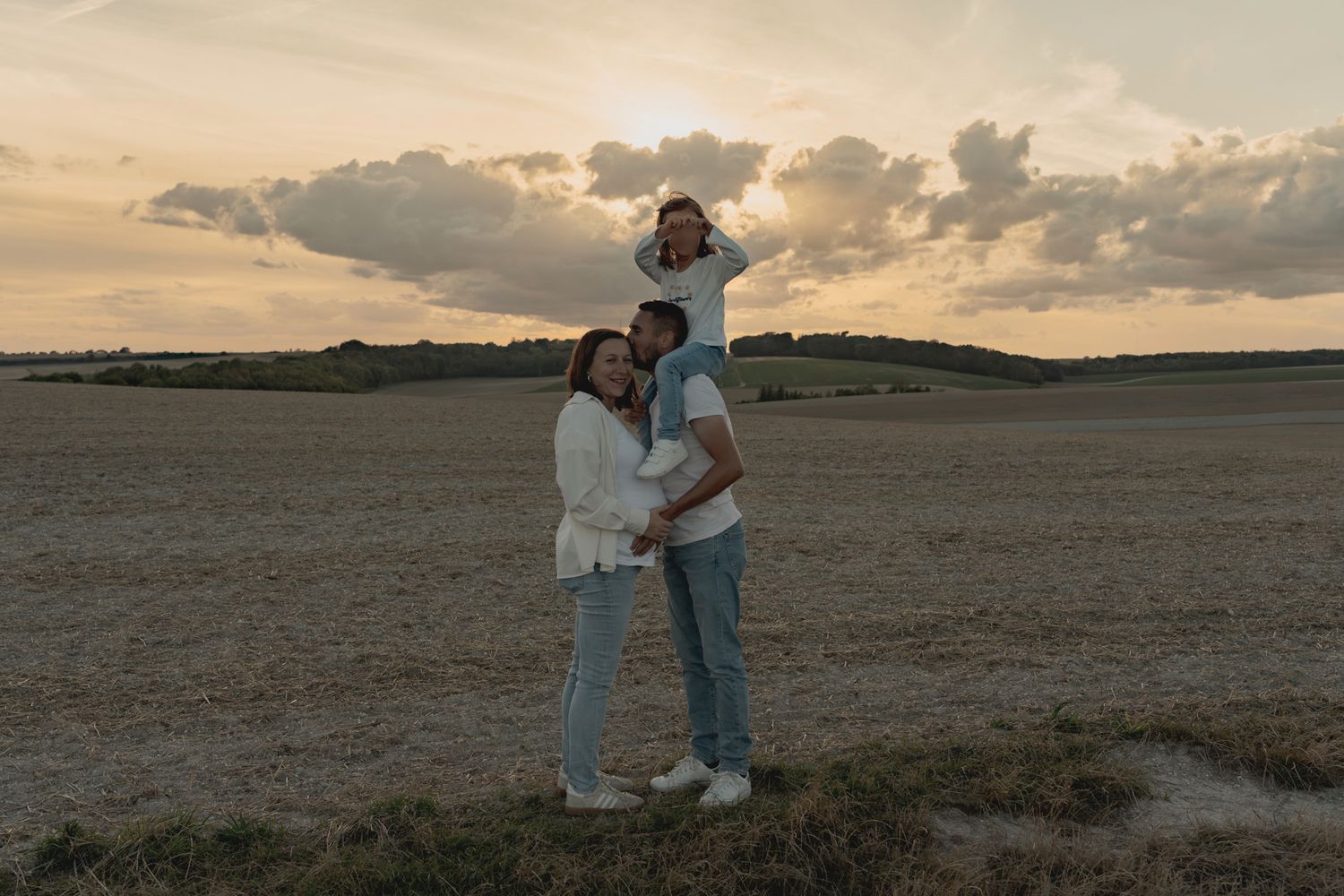 Family of three posing in a golden field at sunset, parents standing with child on shoulders.