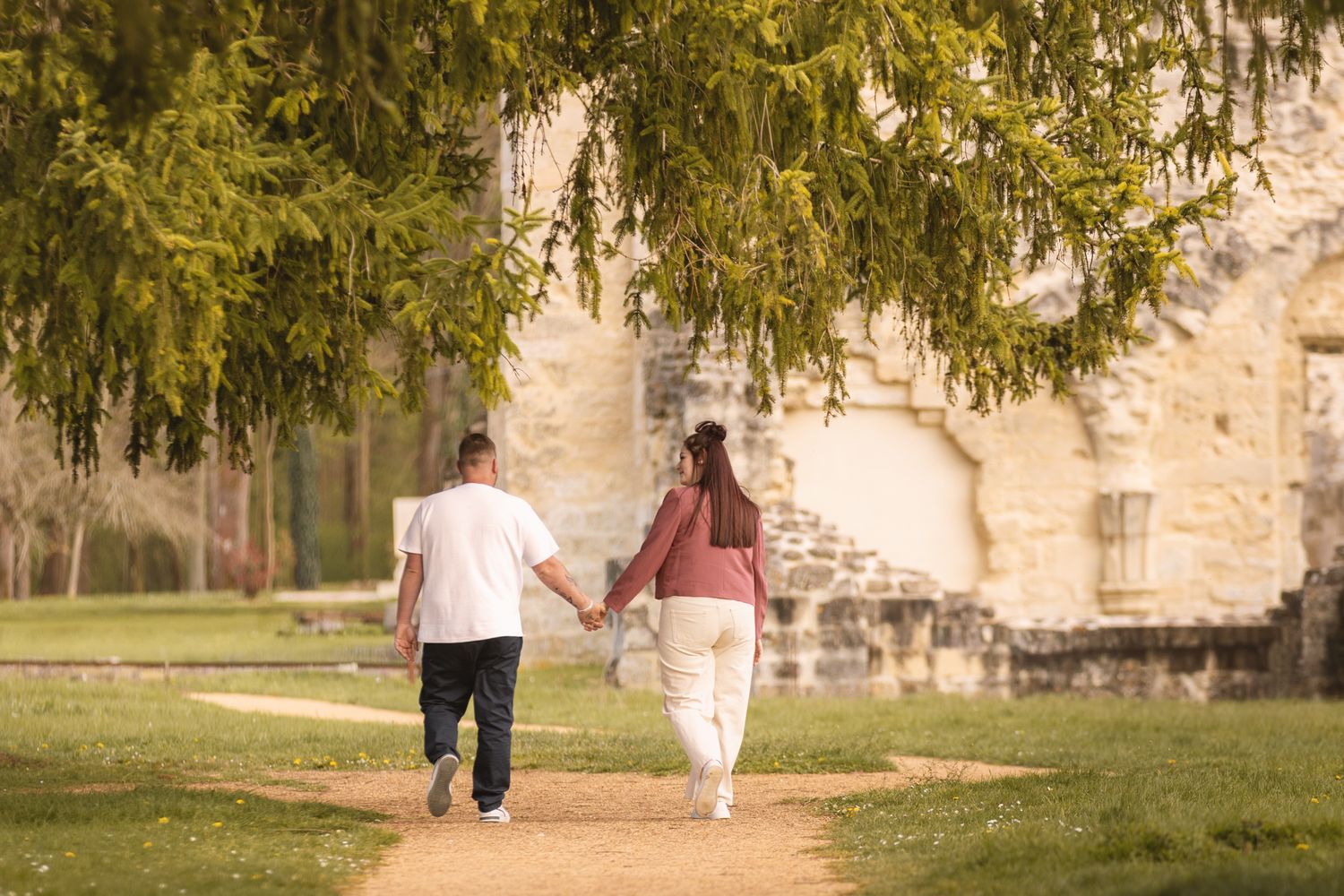 Couple holding hands walking along a garden path beside ancient stone ruins under weeping willow branches.