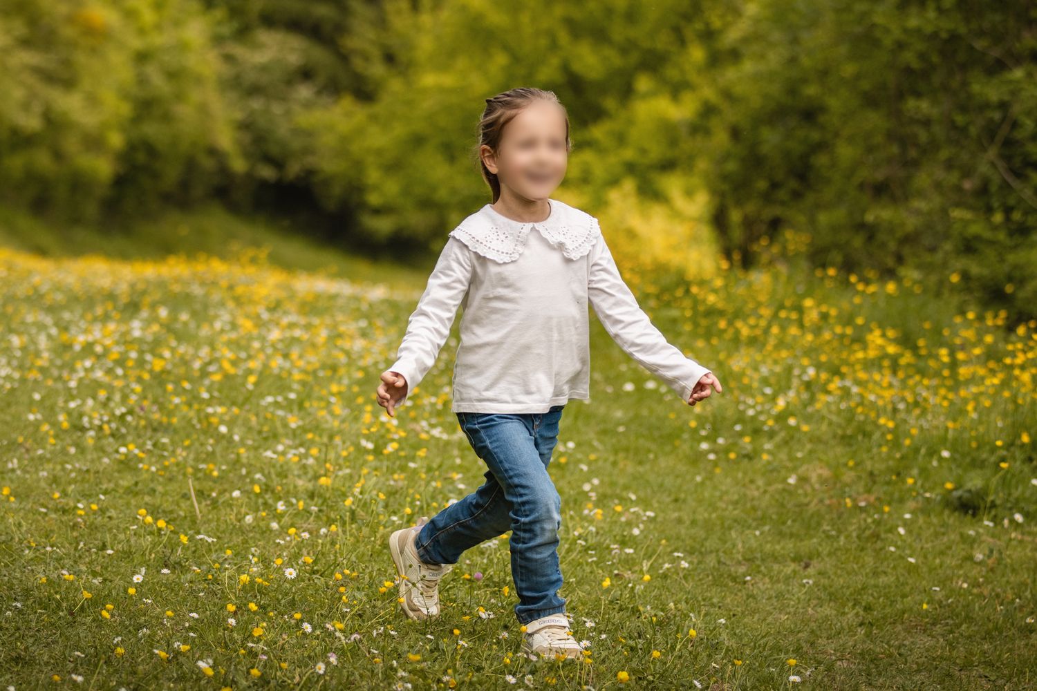 Young girl in white top and jeans running through a sunny wildflower meadow with green trees in background.
