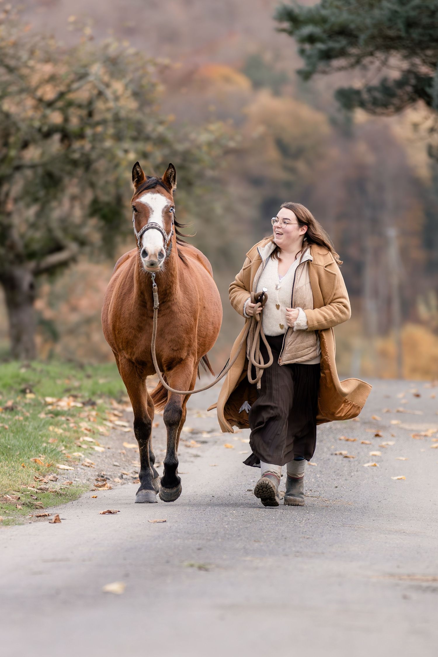 A smiling woman in a tan coat jogs alongside a chestnut horse on a tree-lined autumn road.
