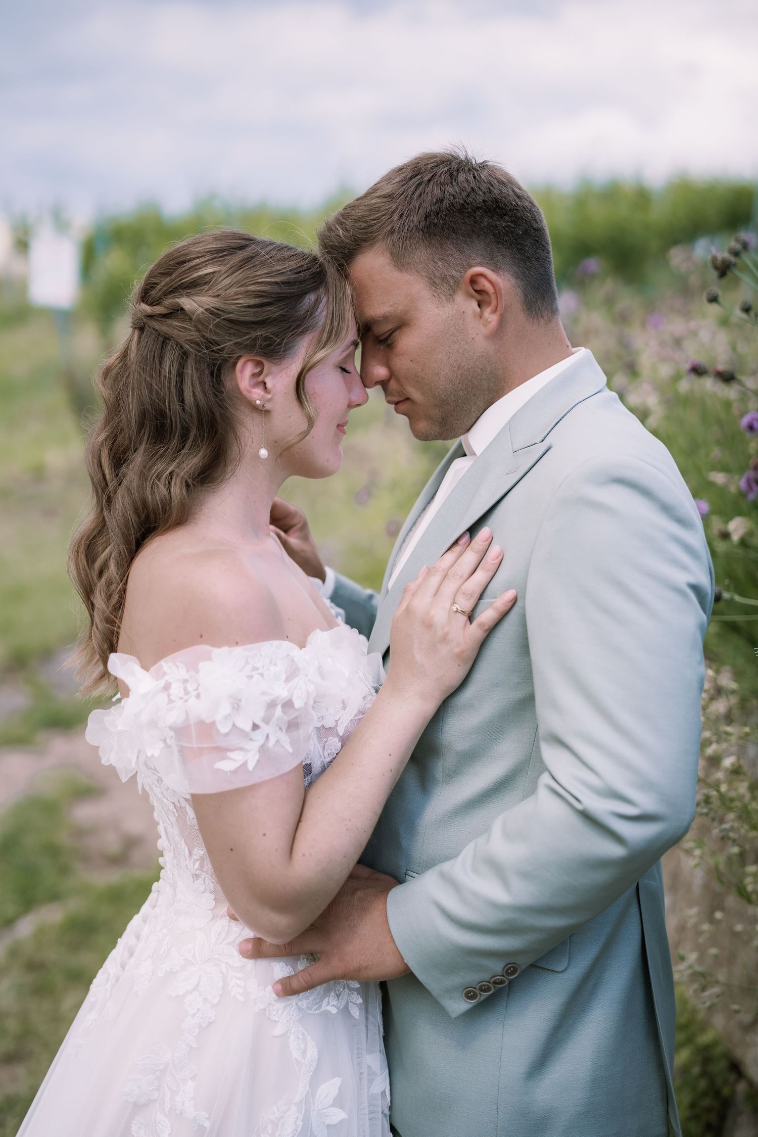 A bride in a white off-shoulder gown and groom in a light blue suit share a romantic moment outdoors.