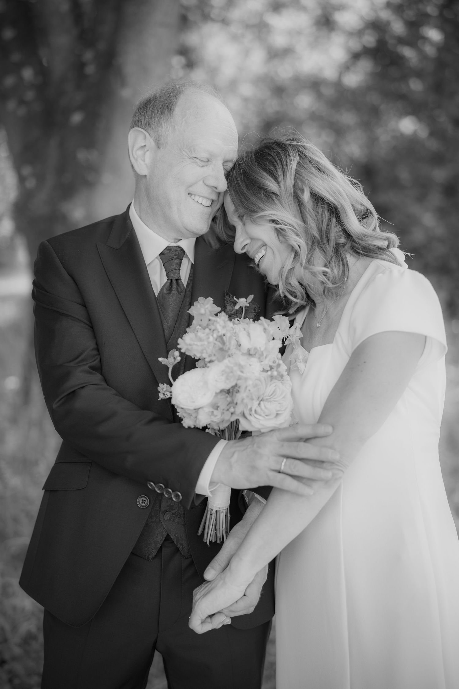 Black and white wedding photo of a smiling couple holding a floral bouquet, sharing a joyful moment outdoors.
