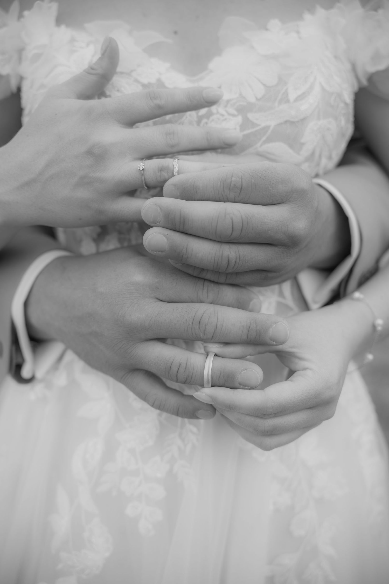 Black and white photo of newlyweds hands with wedding rings, groom embracing bride in lace dress.