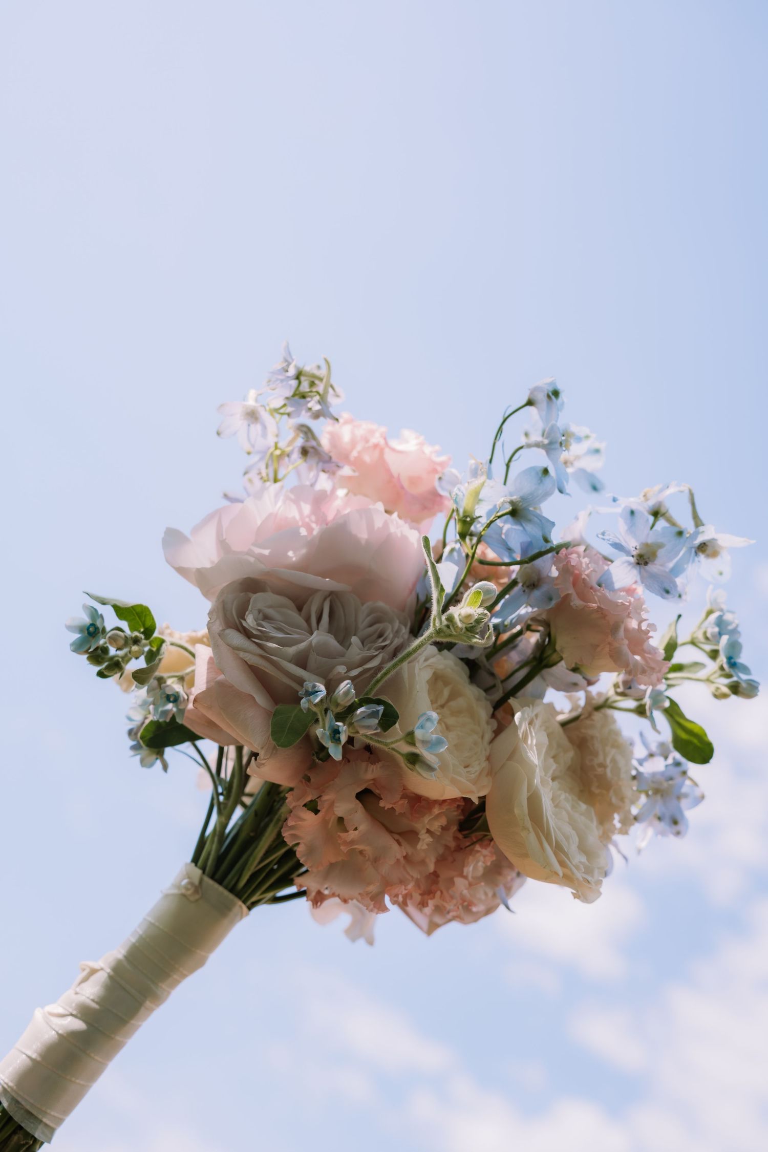 Elegant bridal bouquet of pink roses, white peonies, and blue delphinium held against a clear blue sky.