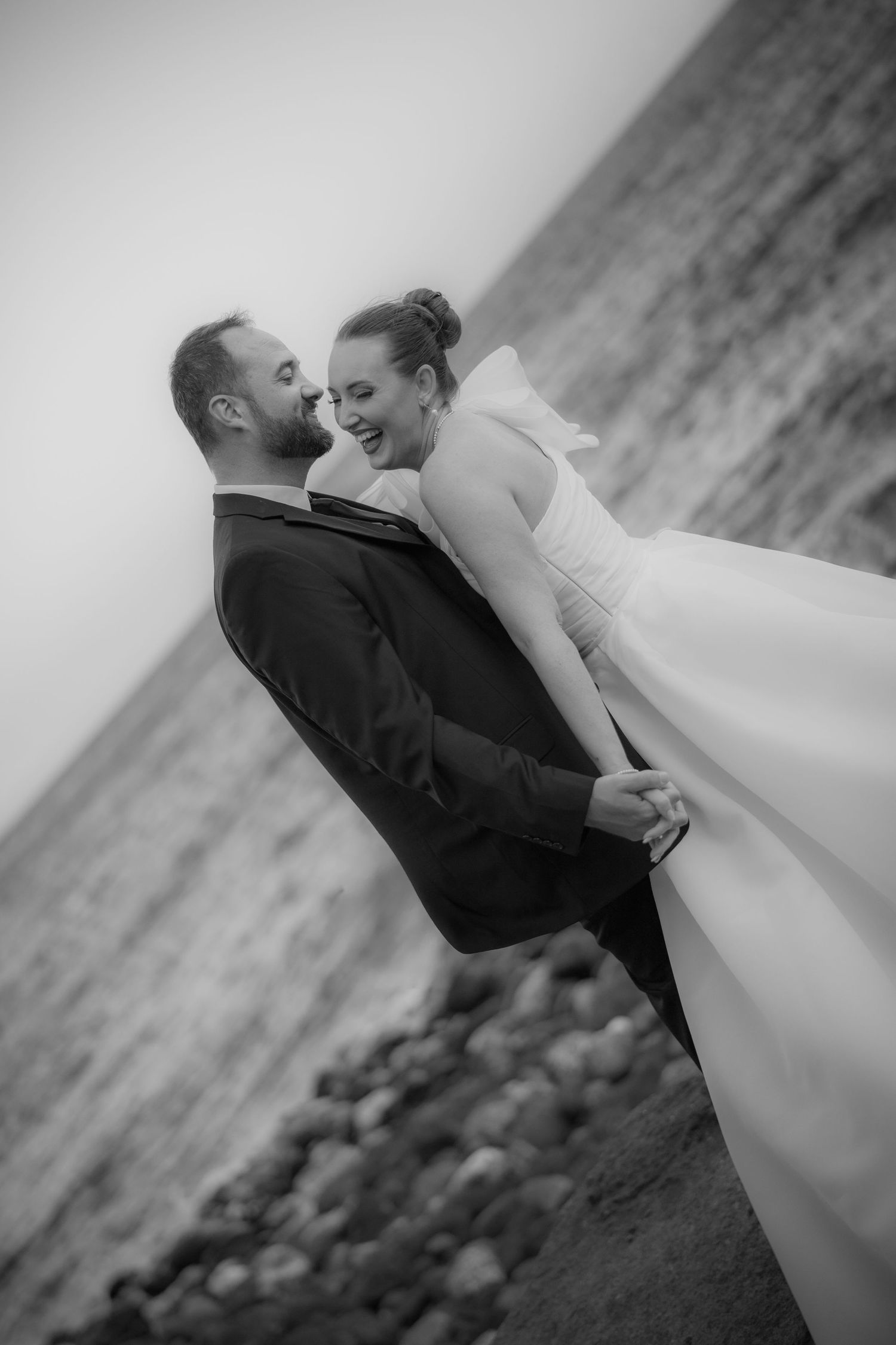 A romantic black and white wedding photo of a couple embracing on a rocky beach with dramatic angle composition.