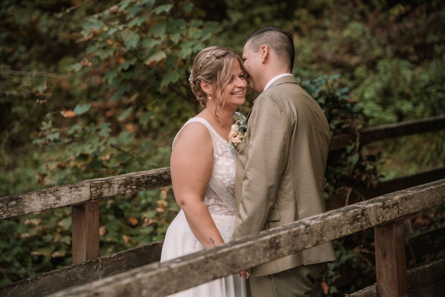 A romantic couple in wedding attire share tender moments on a rustic wooden bridge surrounded by lush greenery.