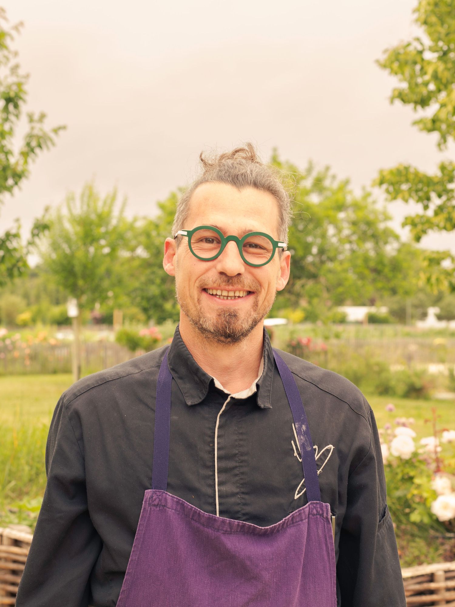Chef portant un tablier violet et des lunettes dans un jardin extérieur.