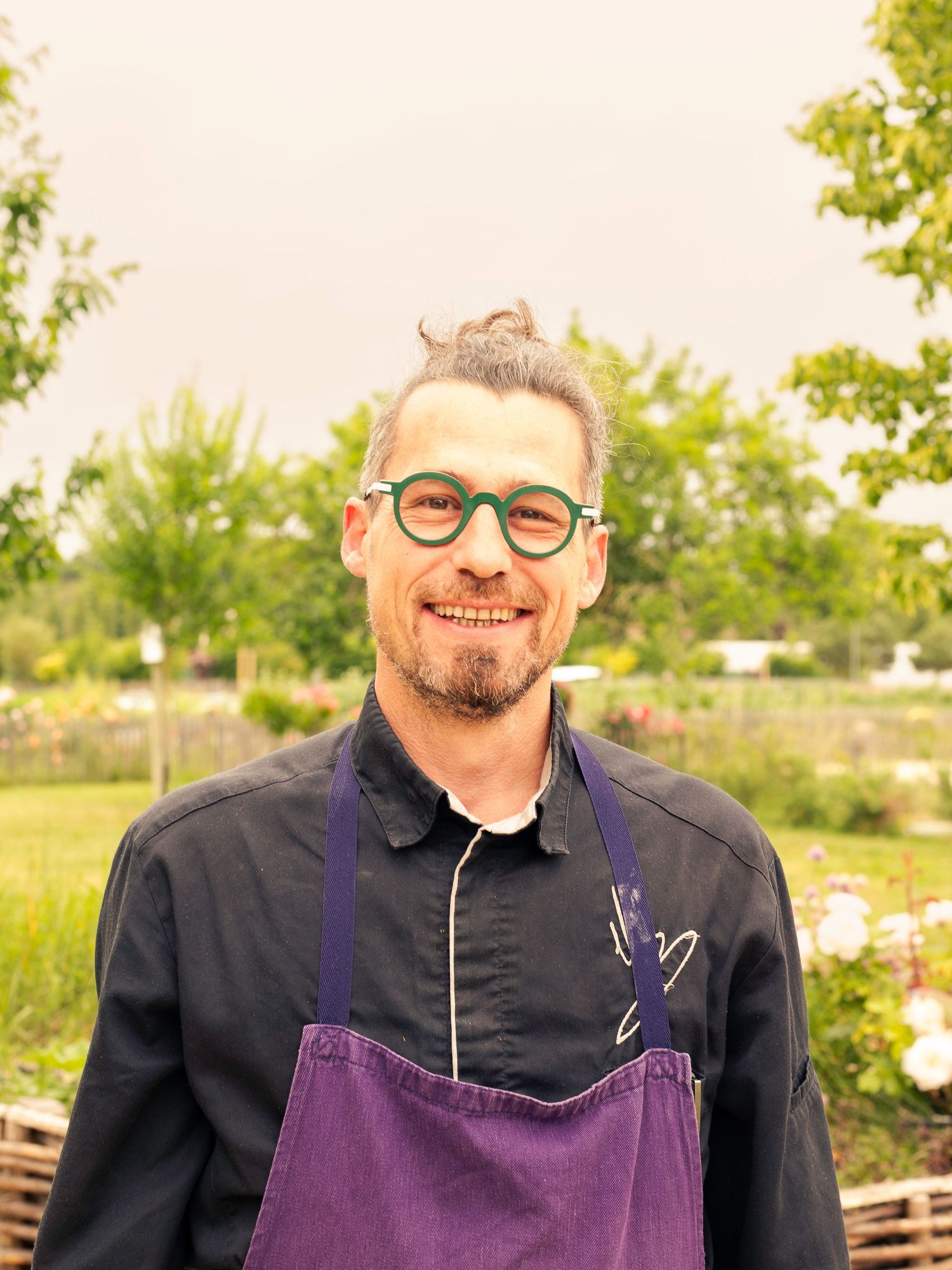 Un chef portant un tablier violet se tient dans un jardin extérieur.