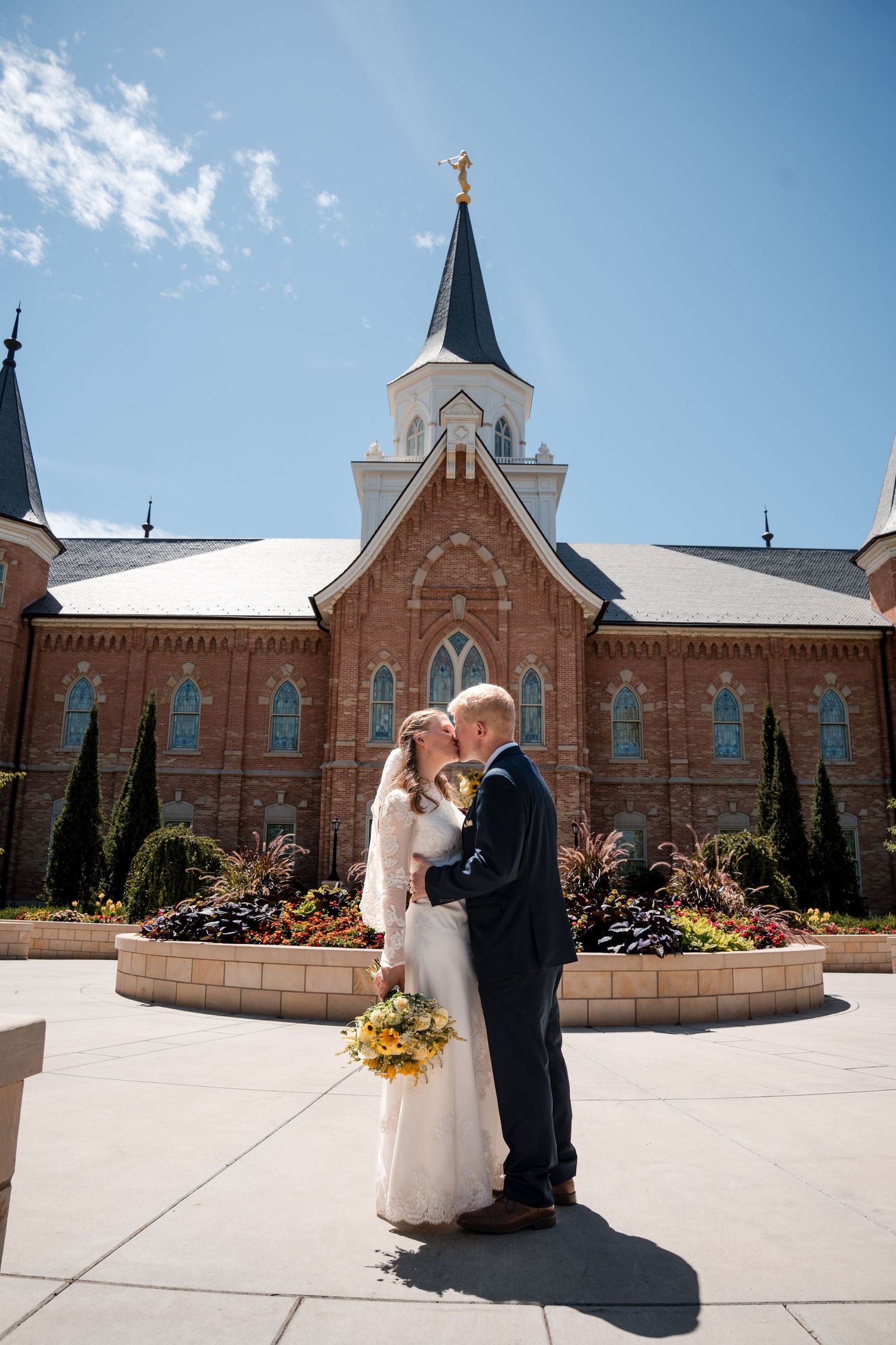 Wedding couple standing in front of the Provo City Center LDS temple with a spire under a bright blue sky.