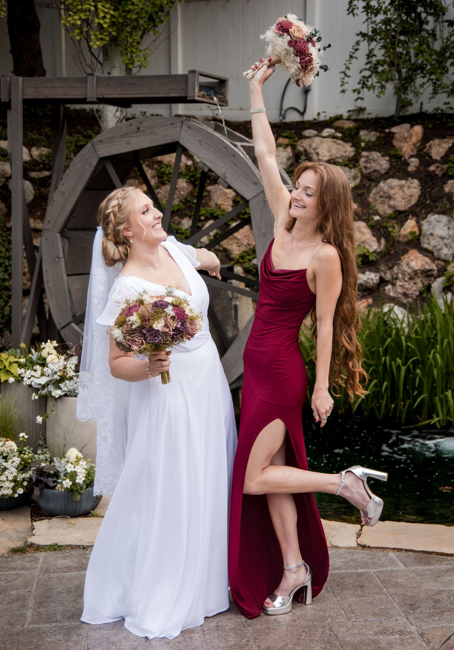 A bride in white and bridesmaid in burgundy celebrate joyfully in a garden setting with bouquets at a wedding.