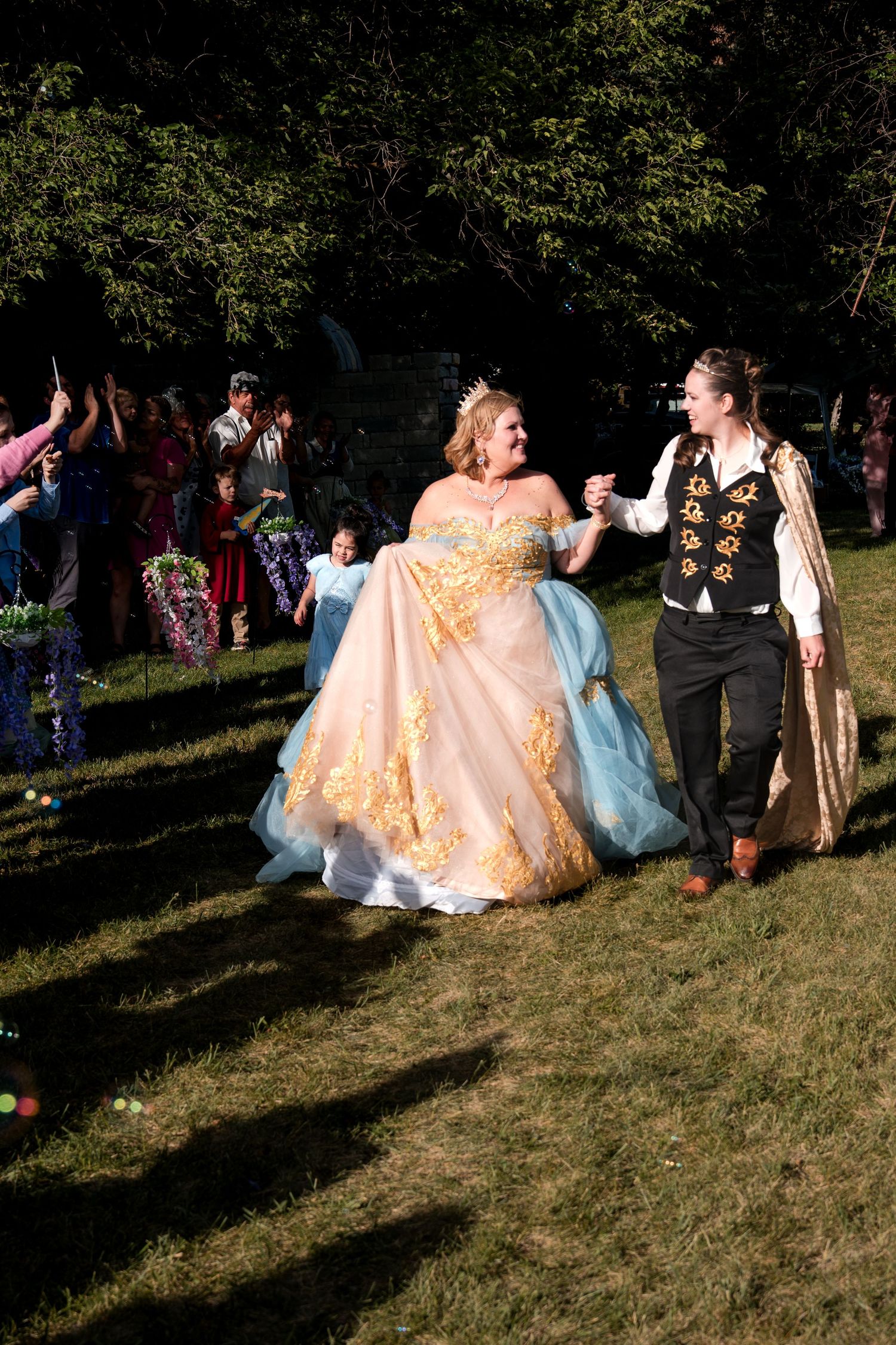 People in elegant ballgowns gather outdoors for a photo session during golden hour at a costumed event in a park setting.