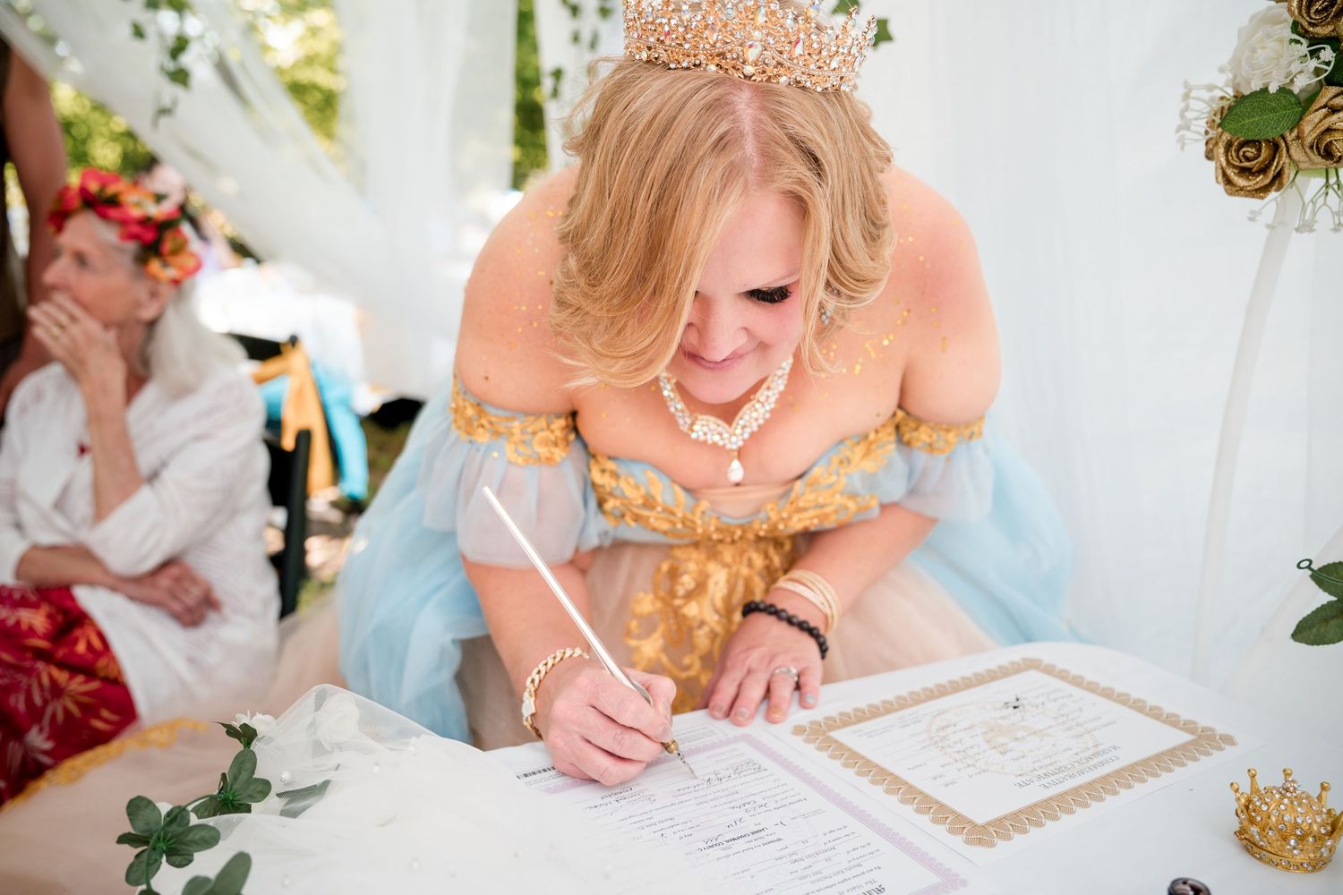 A bride wearing a blue gown and gold crown signs the wedding certificate at a decorated table with floral arrangements.