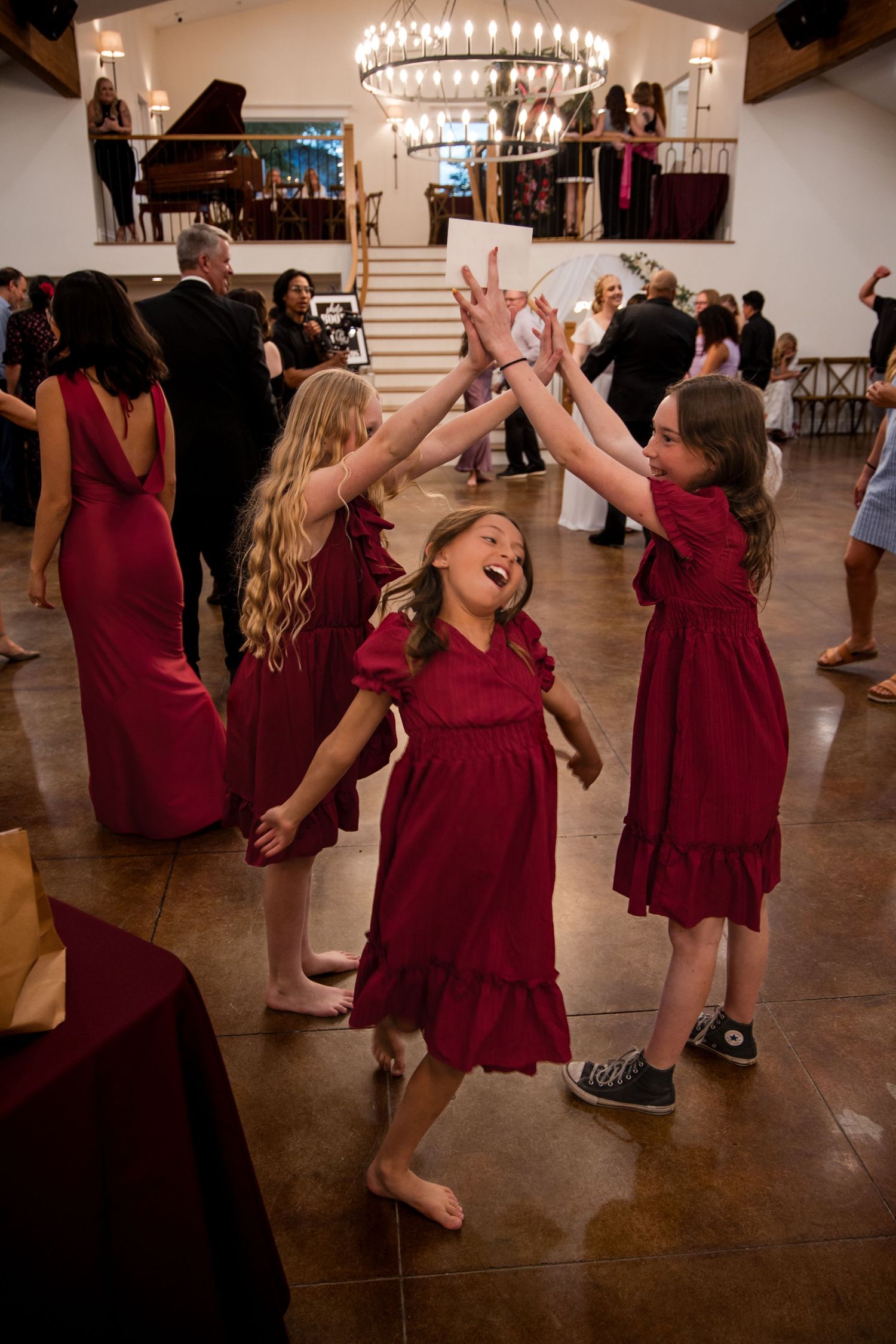 Wedding guests in burgundy dresses dance enthusiastically on a wooden dance floor under chandelier lighting.