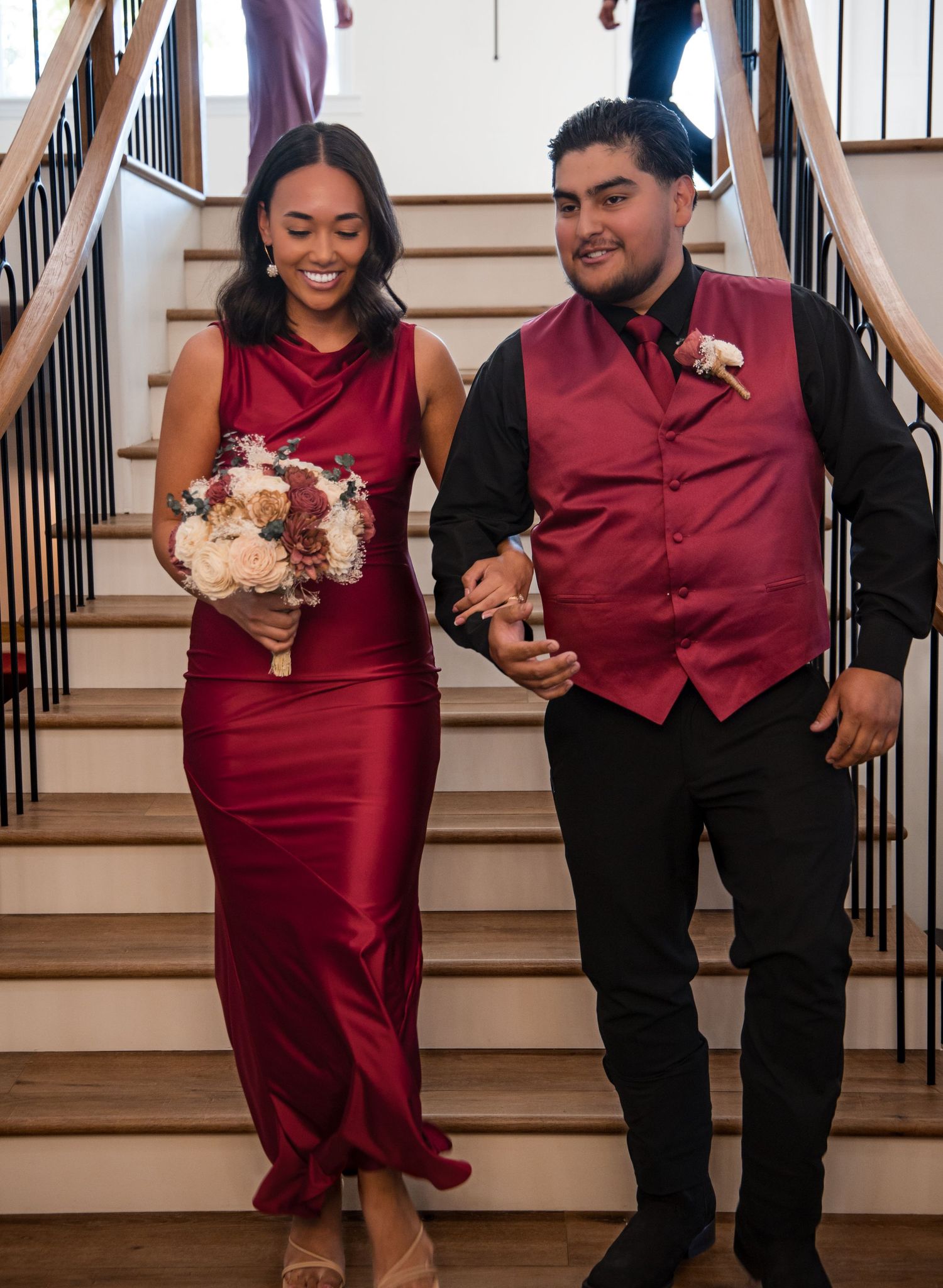 A couple dressed in burgundy formal attire walk down stairs together at a wedding celebration.