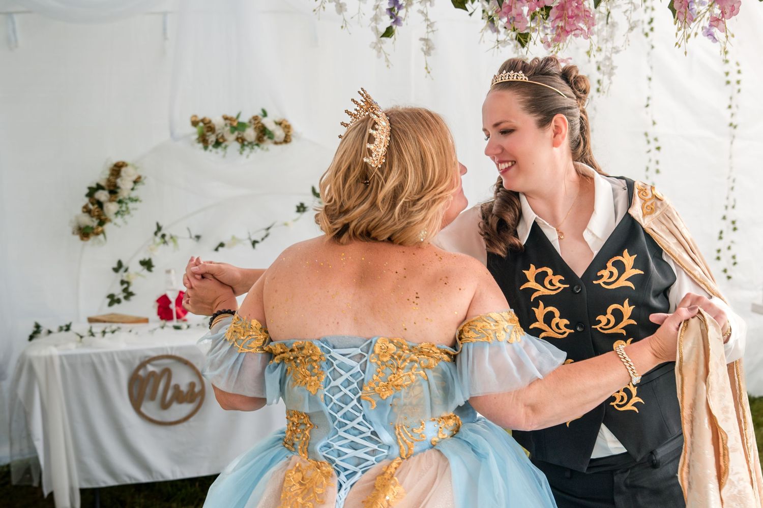 Two brides share a joyful moment at their wedding, one wearing a blue corset ballgown and another in a black floral top.