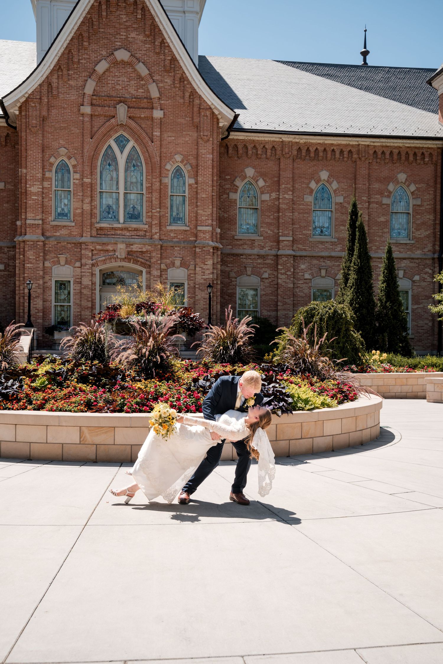 A couple dances in a dip pose outside the Provo City Center LDS temple with Gothic windows and beautiful landscaping.