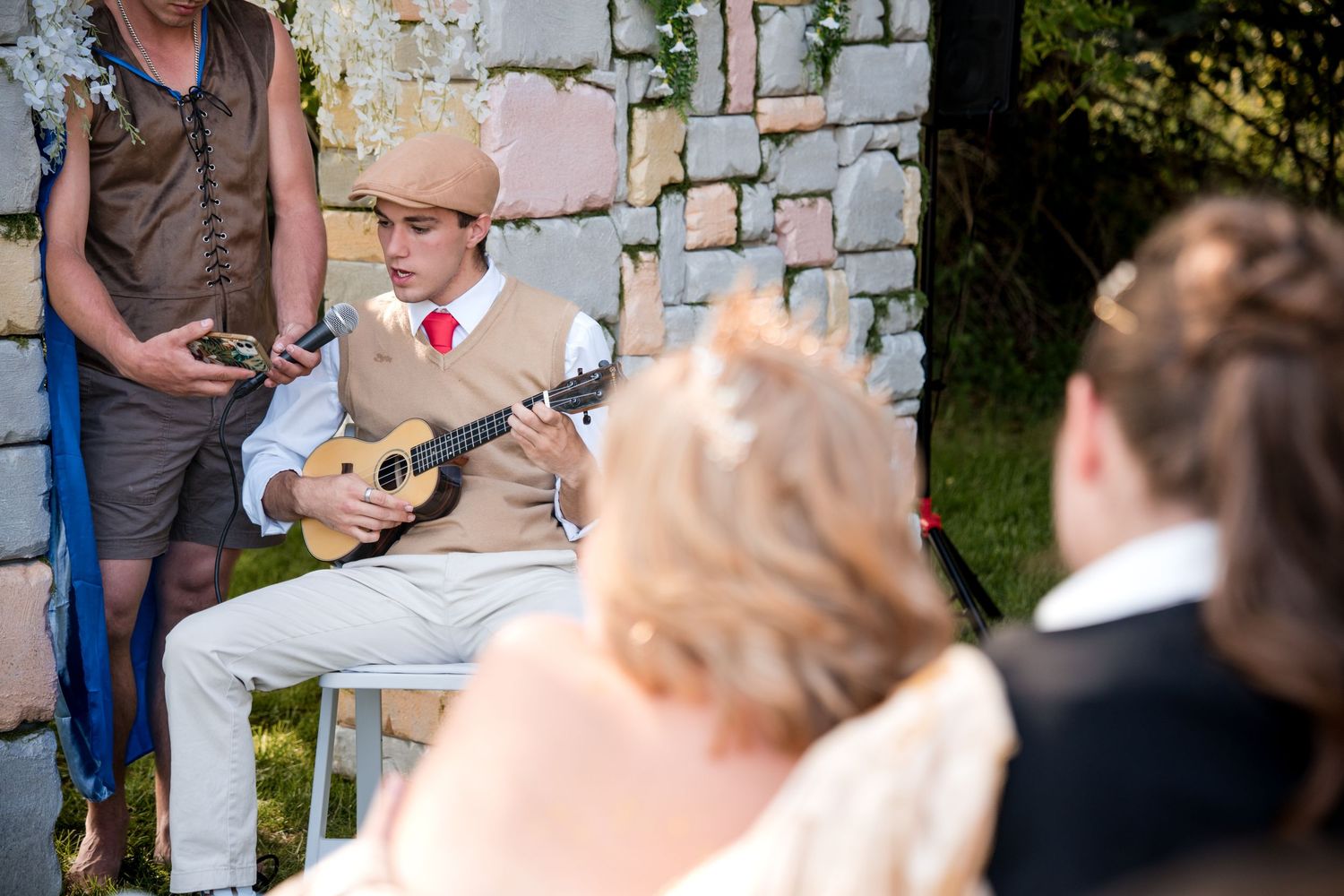 A musician in a light colored suit plays ukulele during a wedding near a stone wall.
