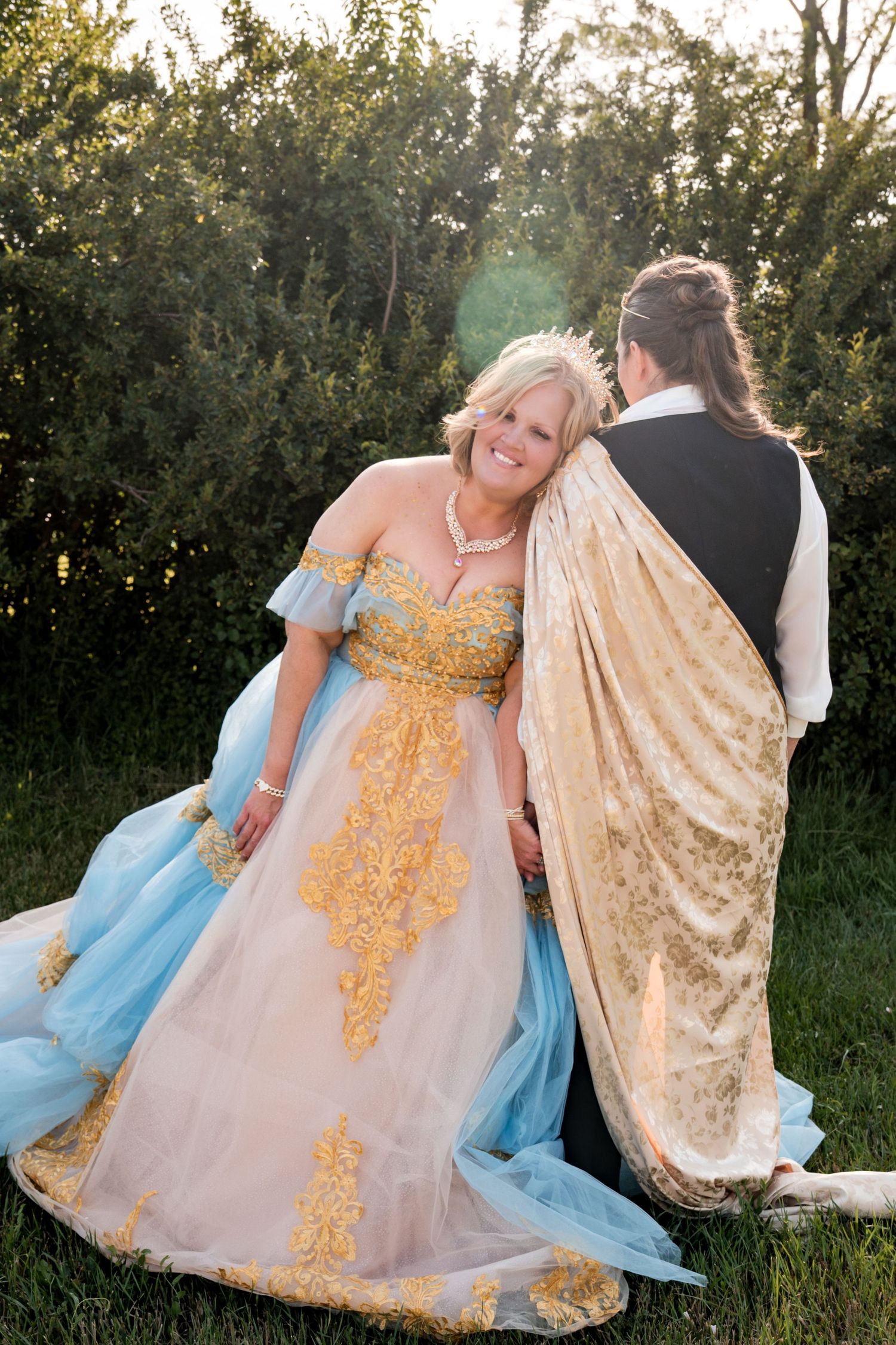 Two people in elegant gowns stand in a sunlit garden, wearing flowy dresses in blue and gold with velvet capes.