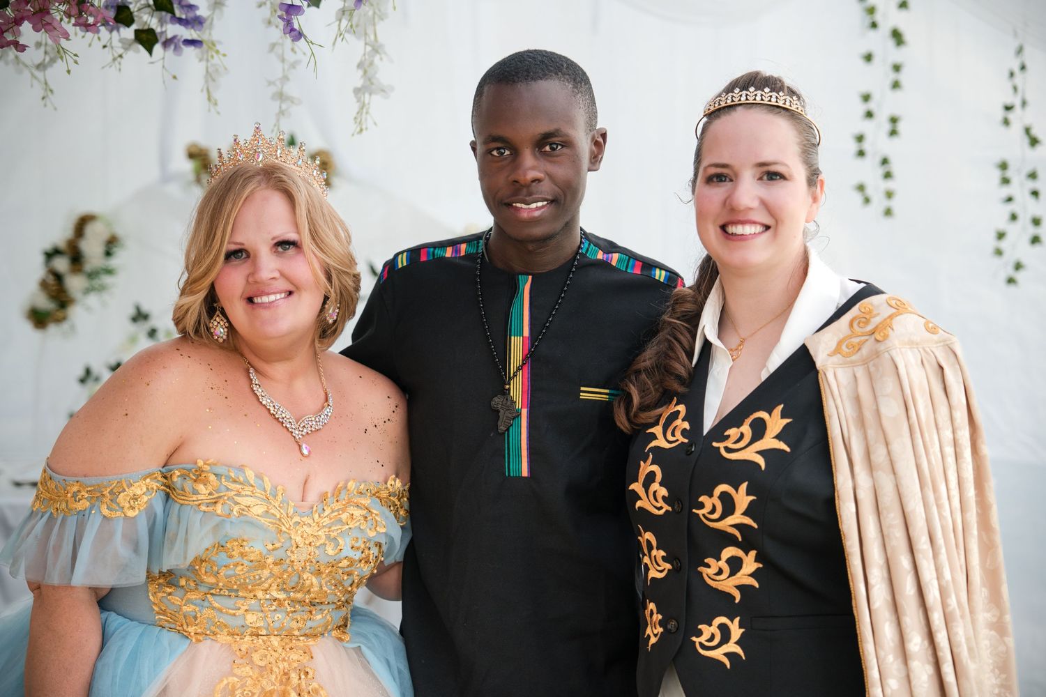 Three people in formal attire pose together at a wedding with floral decorations in the background.