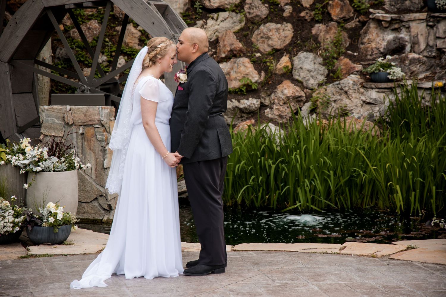 A romantic wedding couple shares a kiss by a stone wall and garden during their outdoor ceremony.