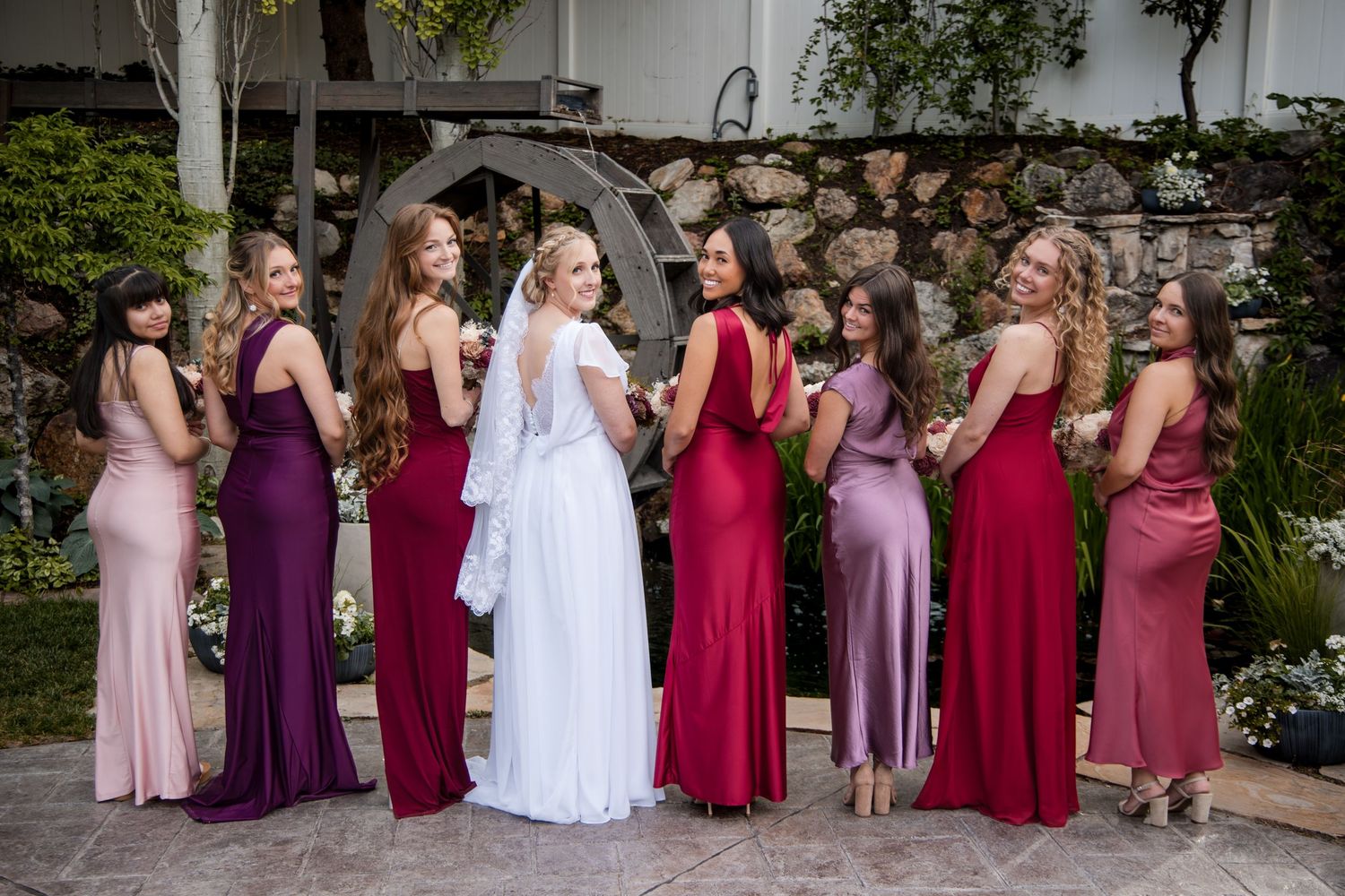 Bridesmaids wearing mix of burgundy and pink floor-length dresses pose with bride in white gown at outdoor garden venue.