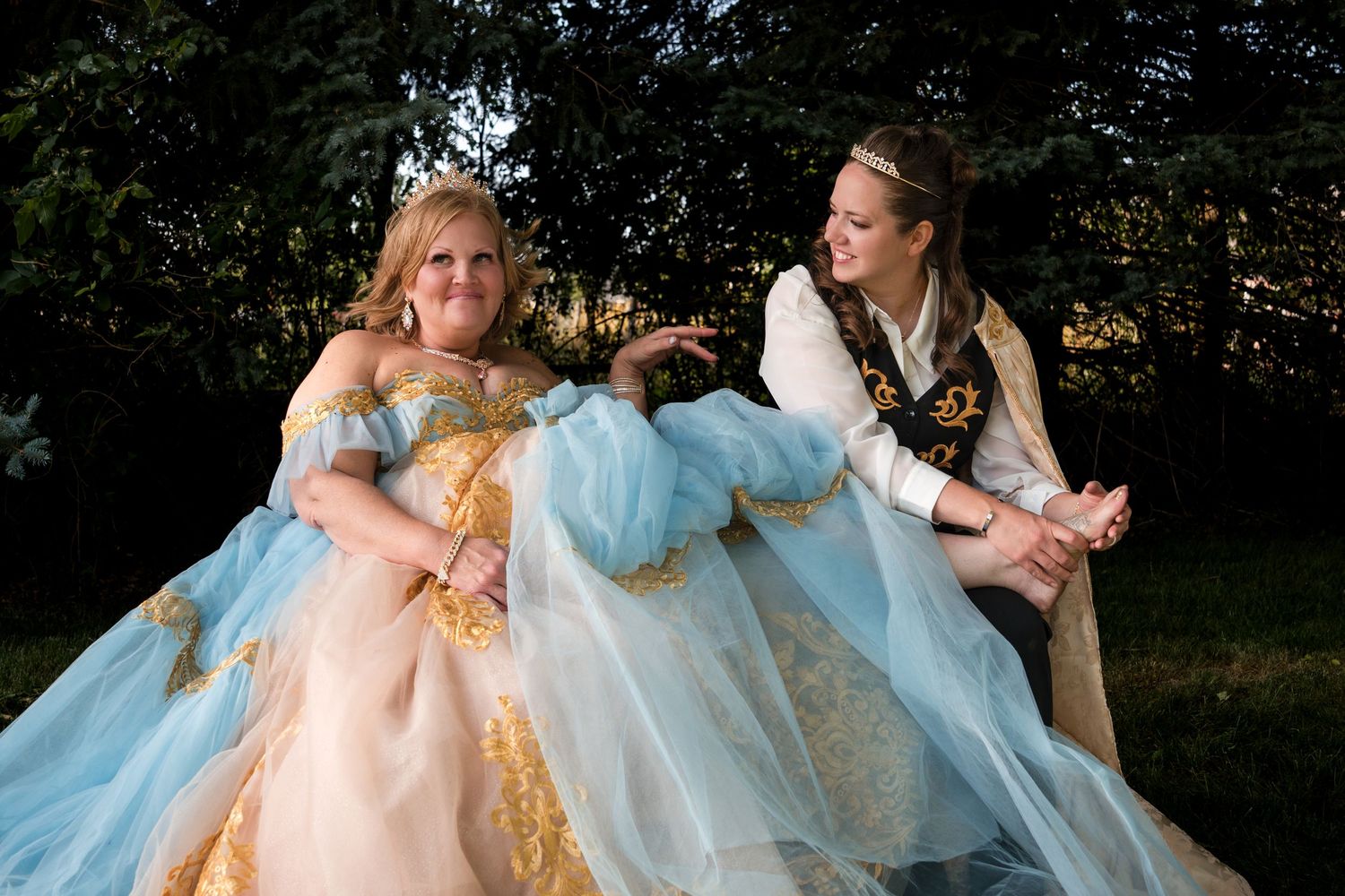 Two people in period costumes share a lighthearted moment while sitting outdoors in elegant dress and pirate attire.