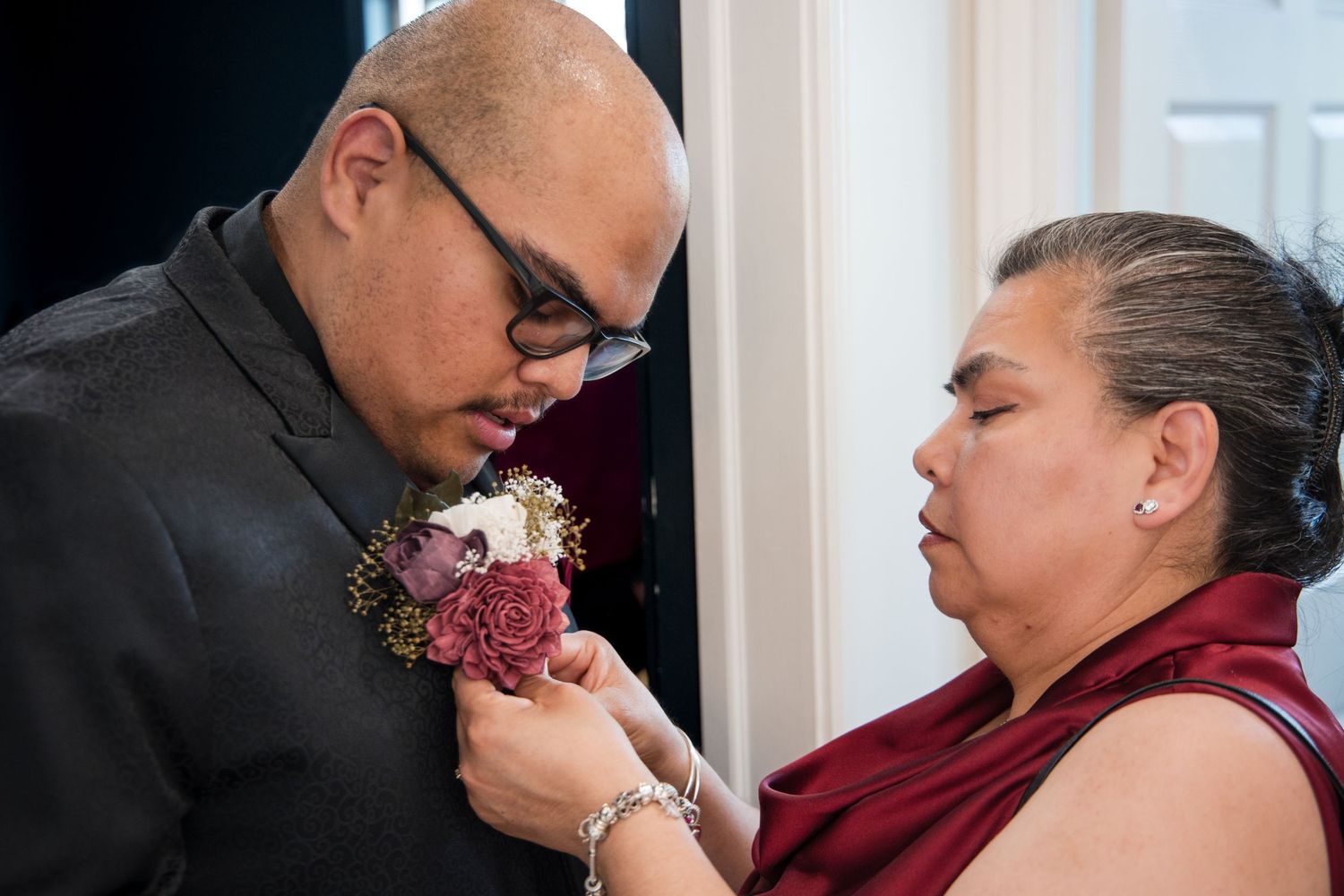 A person in a dark jacket has a pink and purple boutonniere pinned to their lapel by someone wearing burgundy.