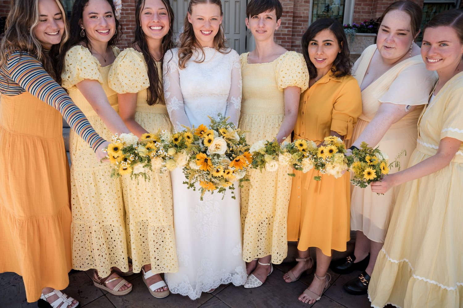 Bridesmaids in coordinated yellow and white dresses pose together holding bright floral bouquets at a wedding event.