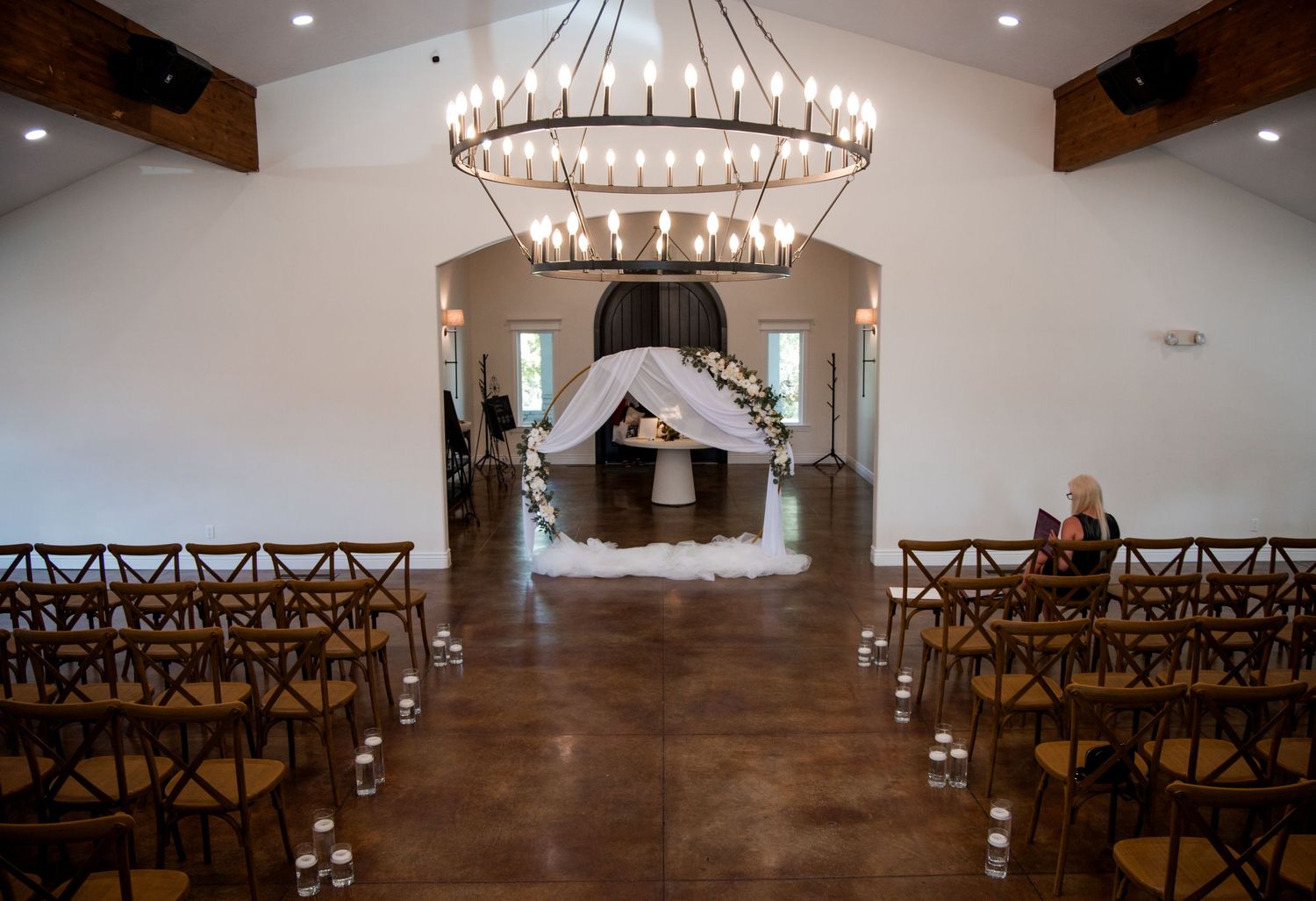 Indoor wedding ceremony space with wooden chairs, circular chandeliers, and a floral arch altar at the end of the aisle.
