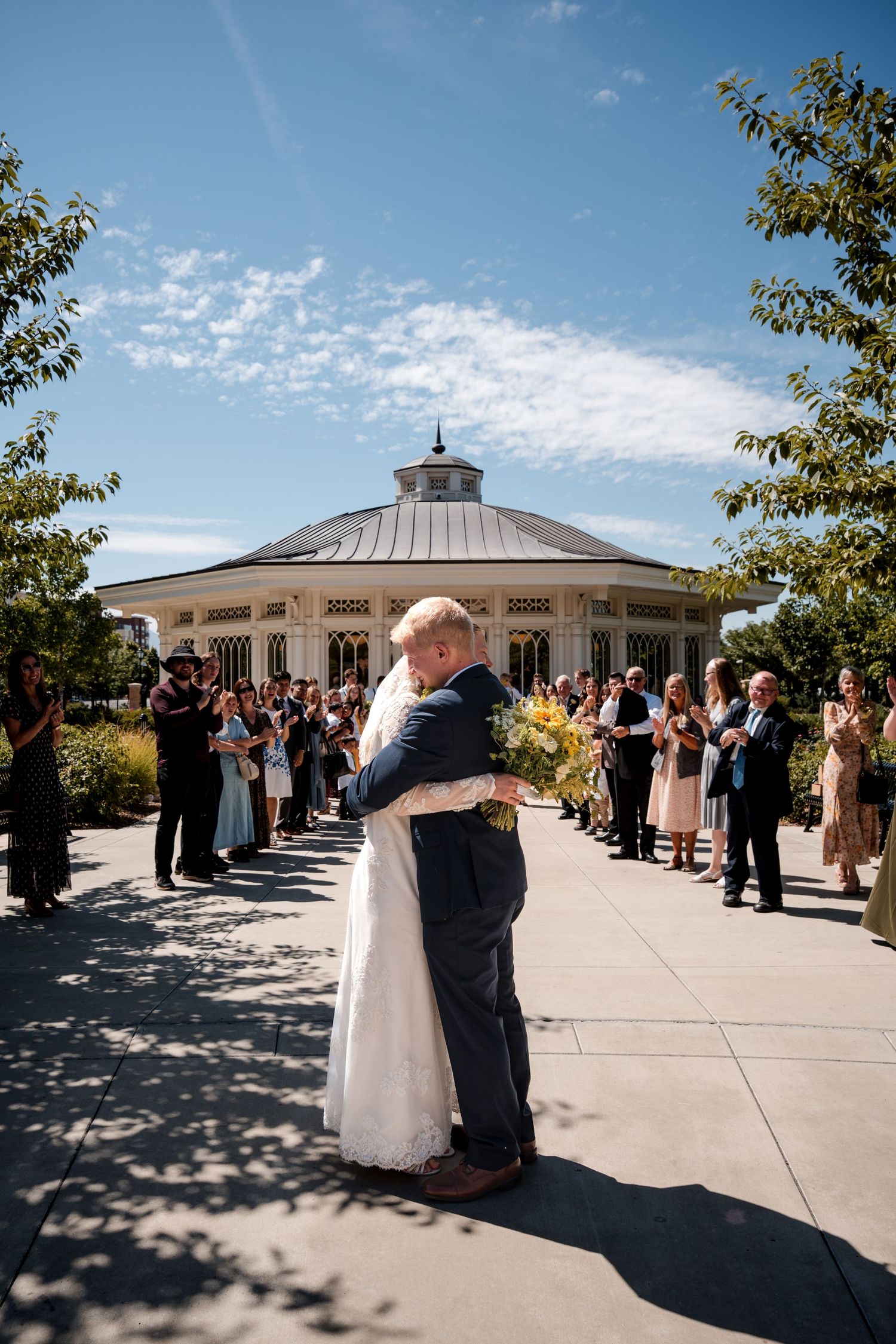 A couple shares their first dance outside a white gazebo reception venue surrounded by wedding guests on a sunny day.