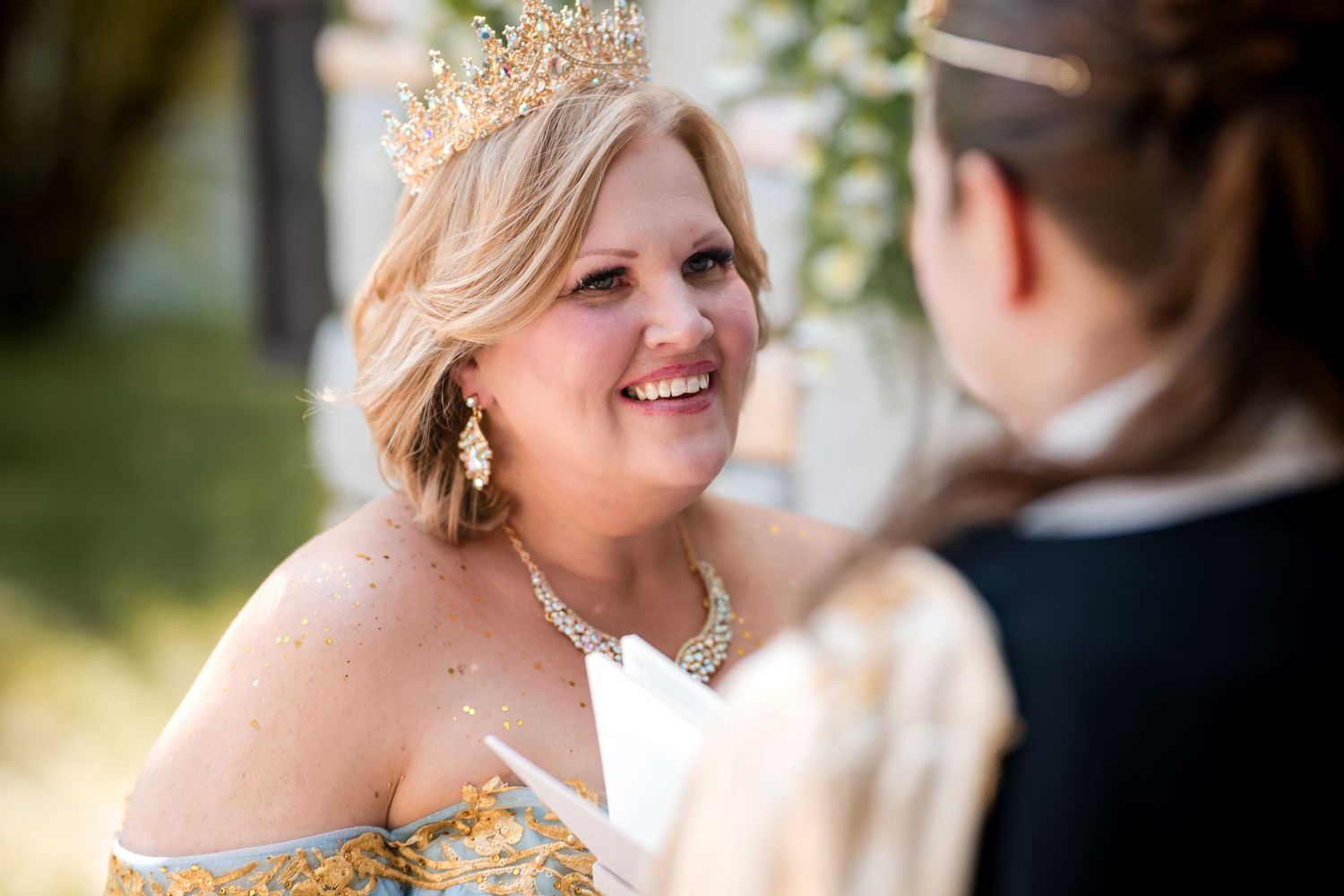 Bride wearing delicate golden crown and strapless dress smiles joyfully during outdoor wedding ceremony.