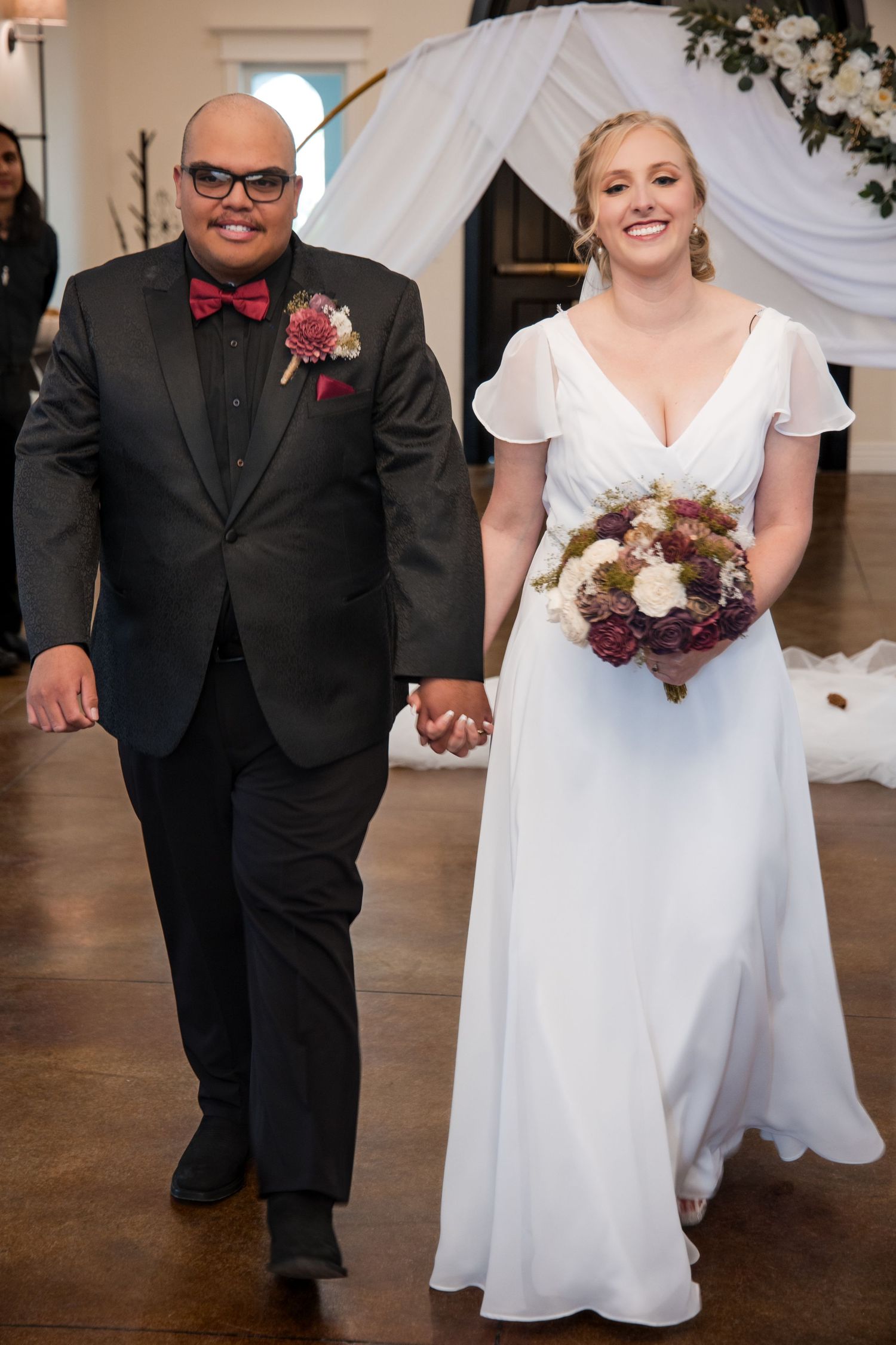 A wedding ceremony at an indoor venue with a white arch, featuring a bride in white gown holding a burgundy bouquet.