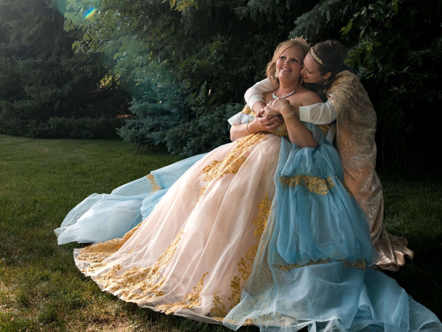 Two people in elegant vintage-style ball gowns share a tender embrace while sitting on grass in a sunlit garden setting.