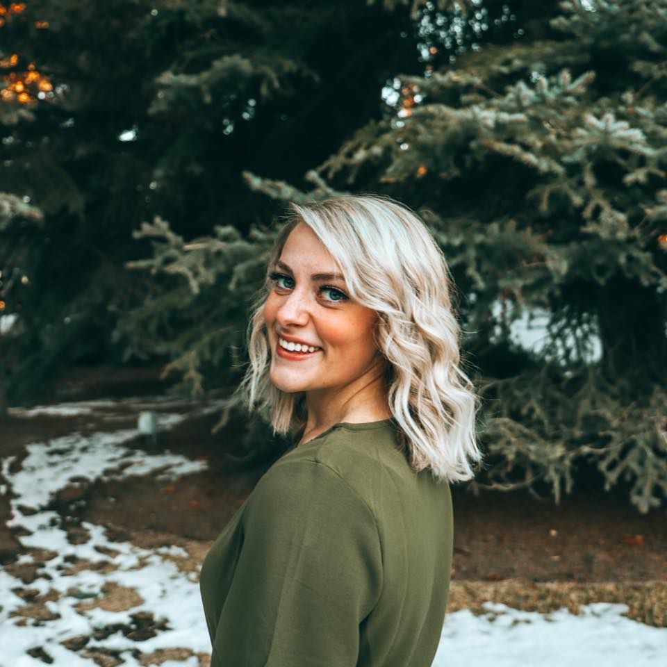 Person in an olive green sweater poses outdoors in a snowy winter setting with evergreen trees in background.