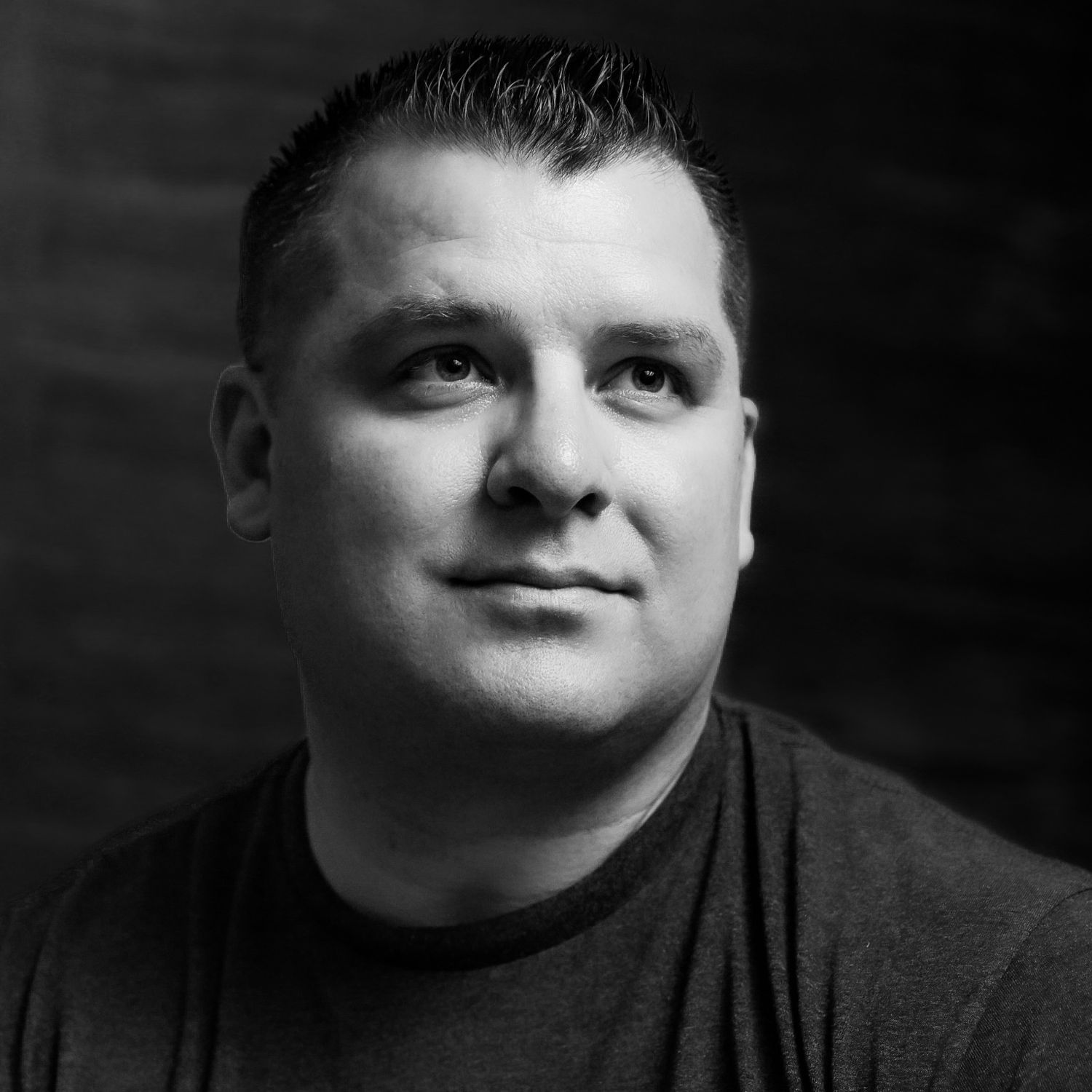Black and white portrait of someone in a dark t-shirt against a dark background with dramatic lighting.
