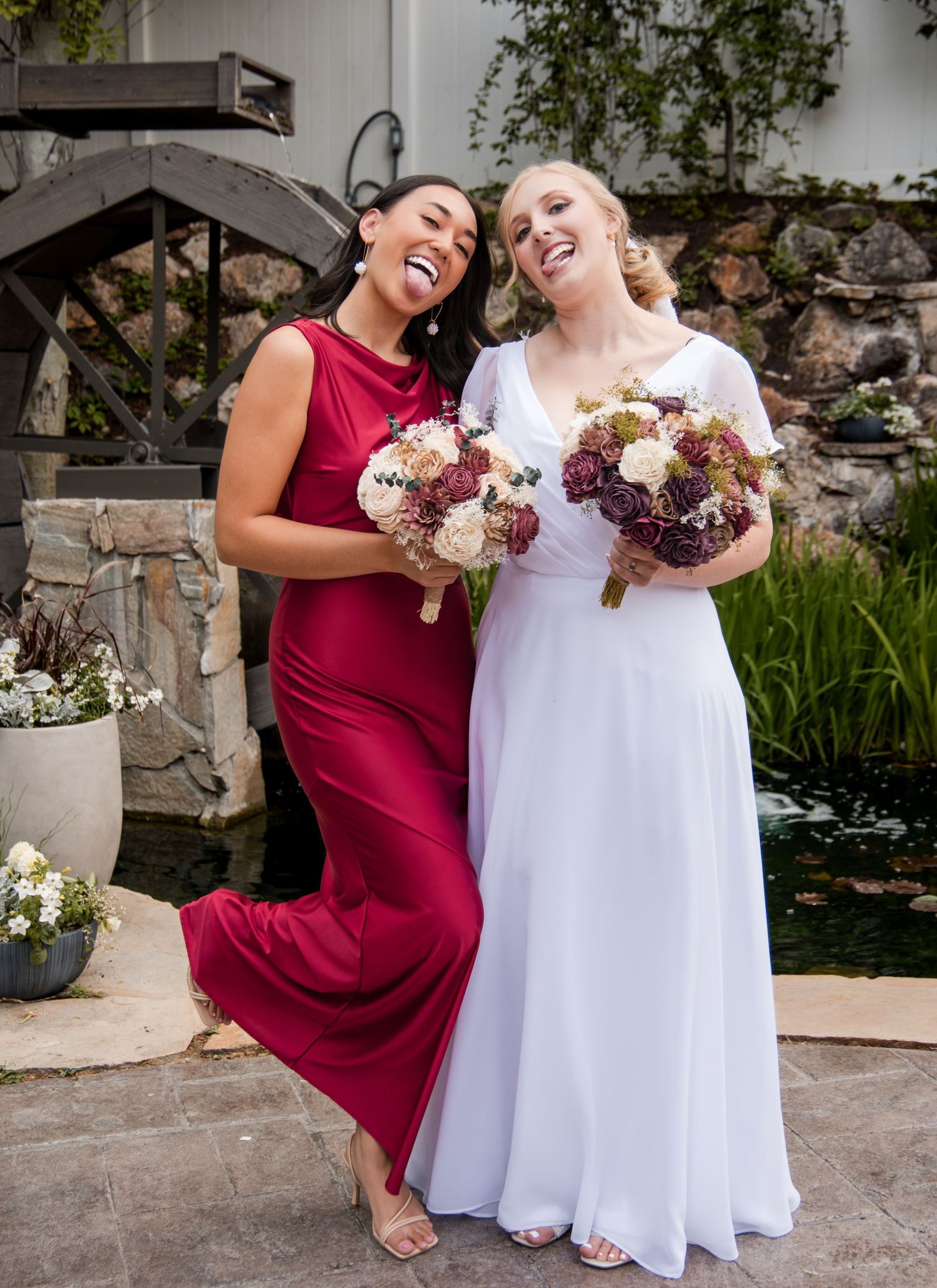 Two wedding attendants in burgundy and white dresses laugh joyfully while holding matching floral bouquets outdoors.