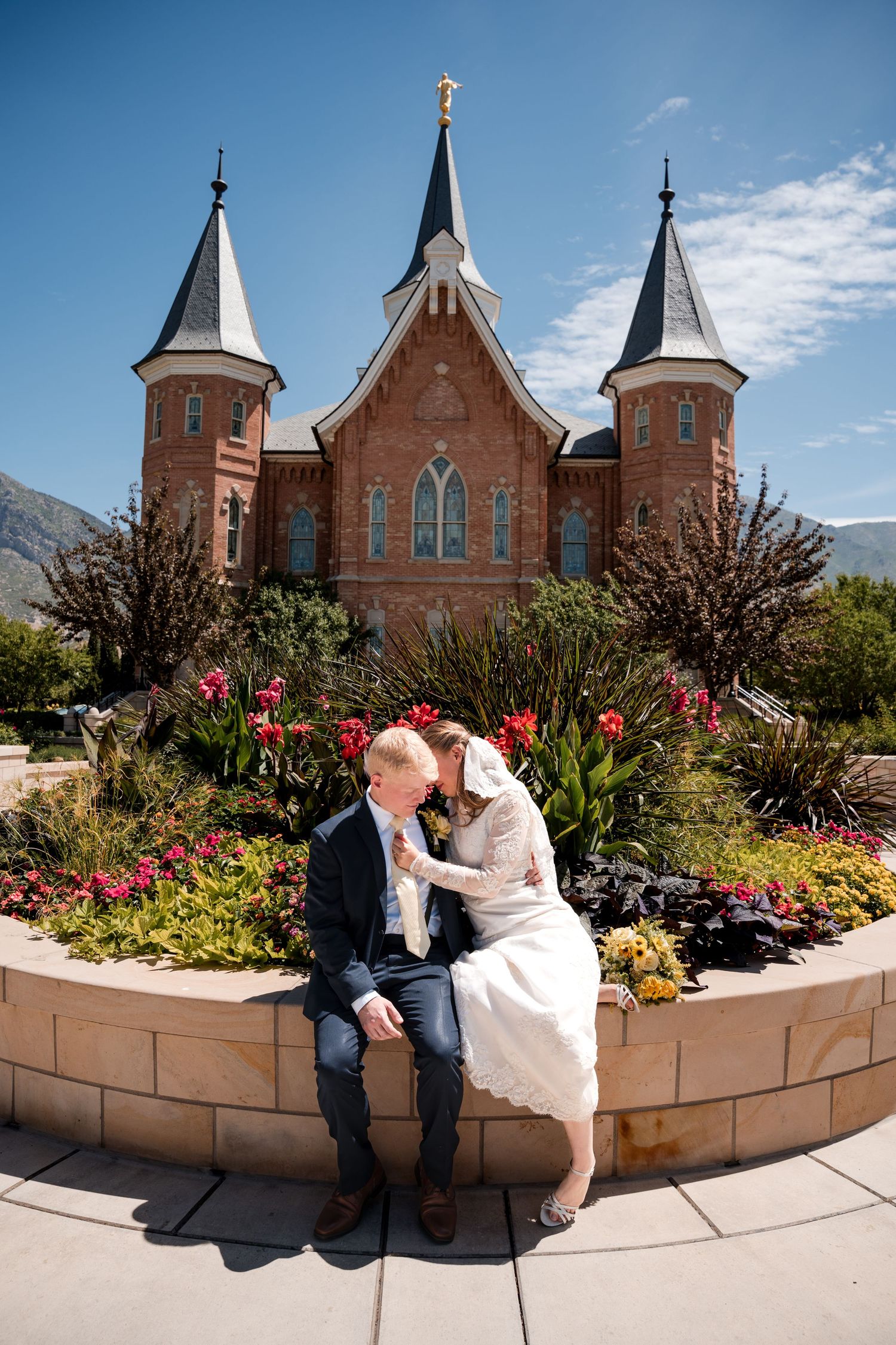 A couple sits in front of Provo City Center LDS temple with spires and red tulips blooming in a garden on a sunny day.
